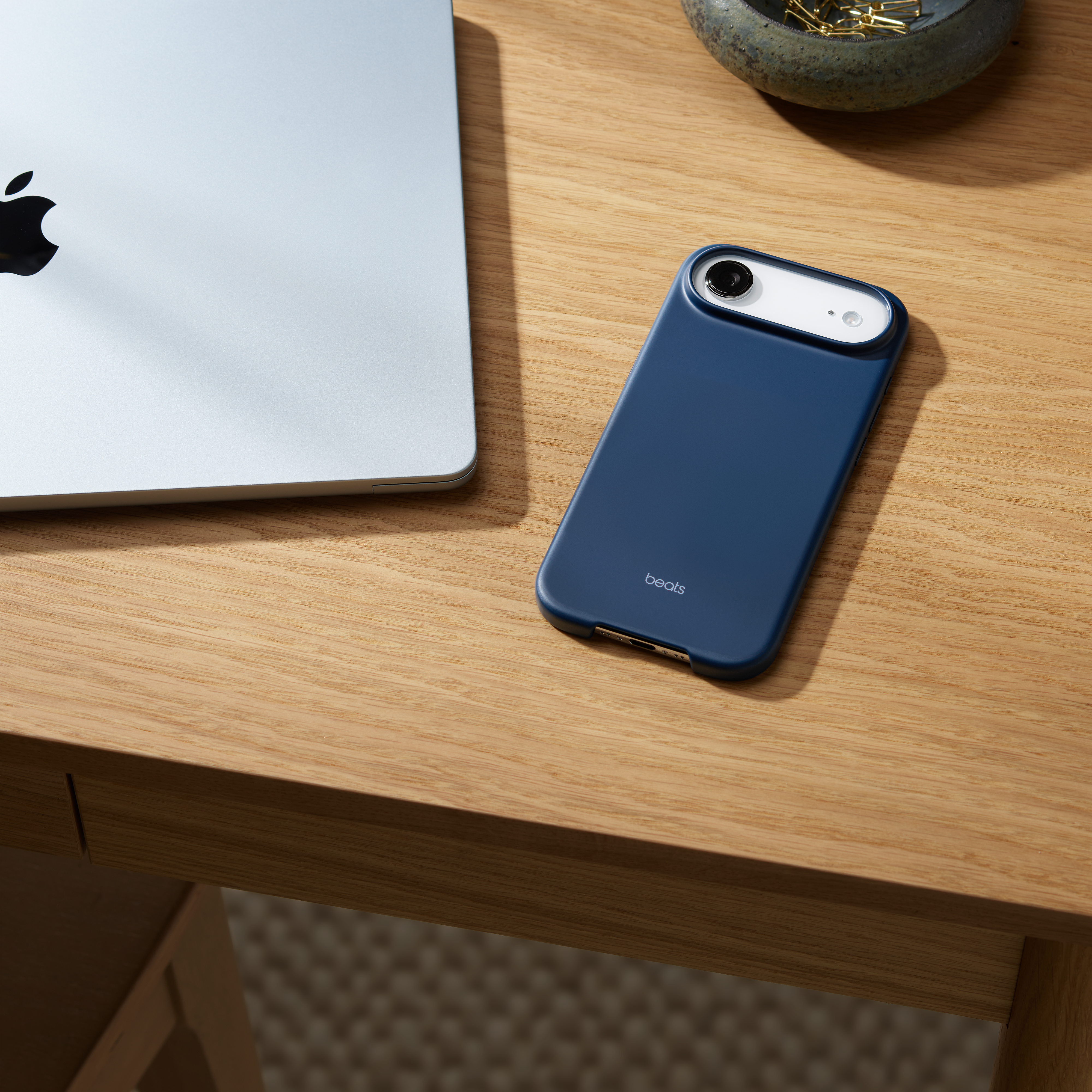 Un étui de téléphone bleu avec 'beats' sur un bureau en bois. Un ordinateur portable et un bol avec des trombones dorés sont également présents.