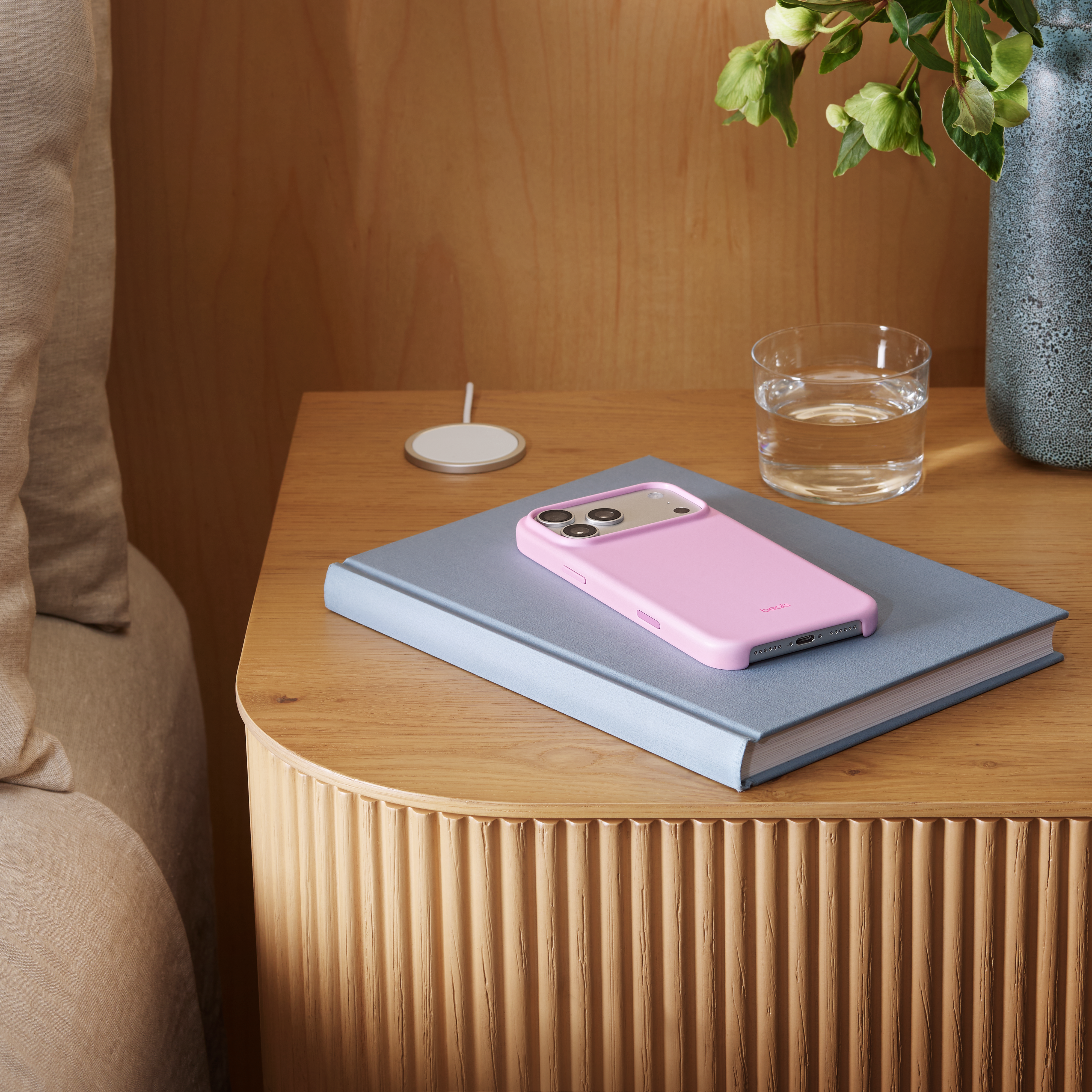 Ein Nachttisch mit Telefon, Ladegerät, Wasserglas und Vase. Es hat eine hellbraune Holzmaserung.