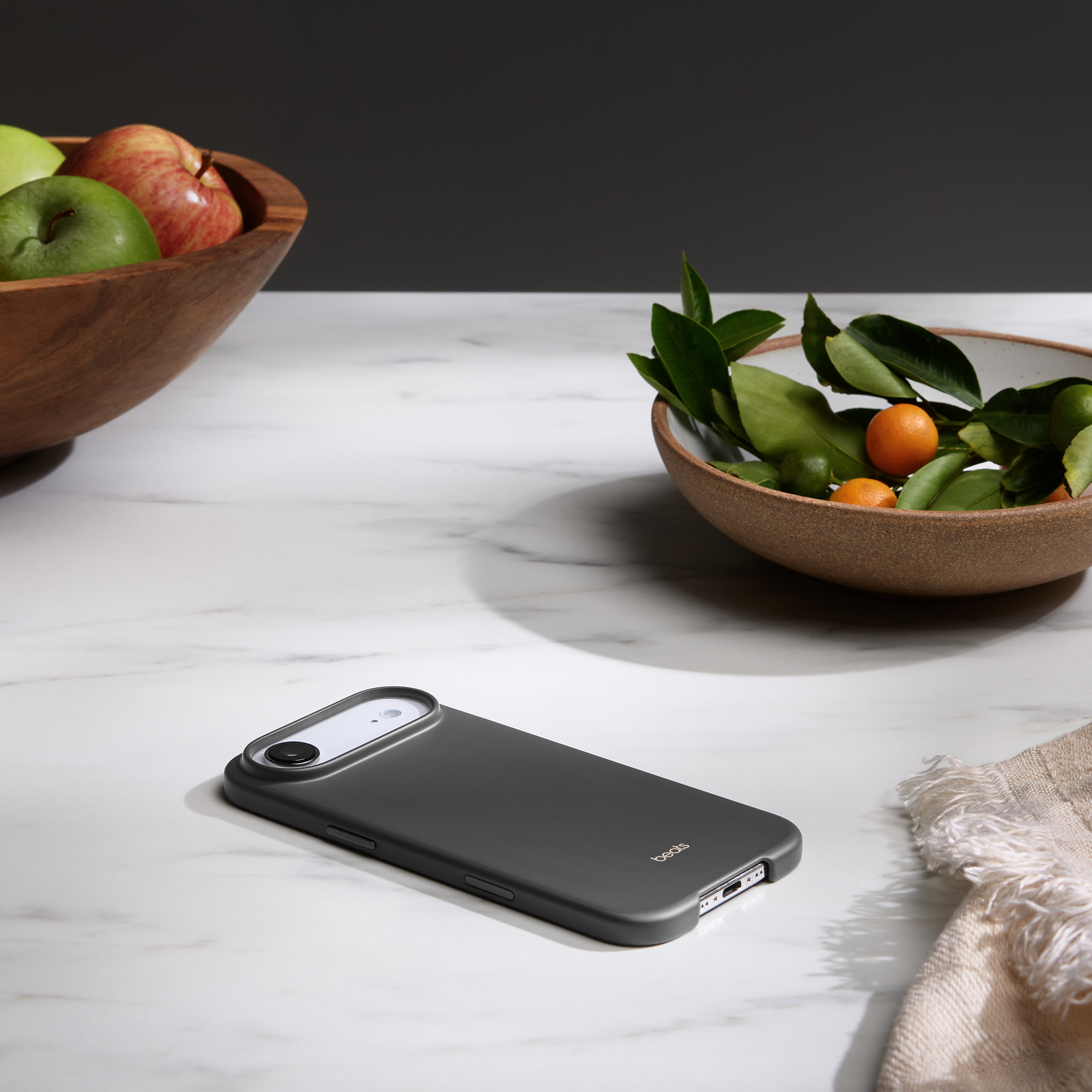 Un étui de téléphone gris est posé sur une surface en marbre. Des bols de fruits et de feuilles sont en arrière-plan. Un tissu beige est à droite.