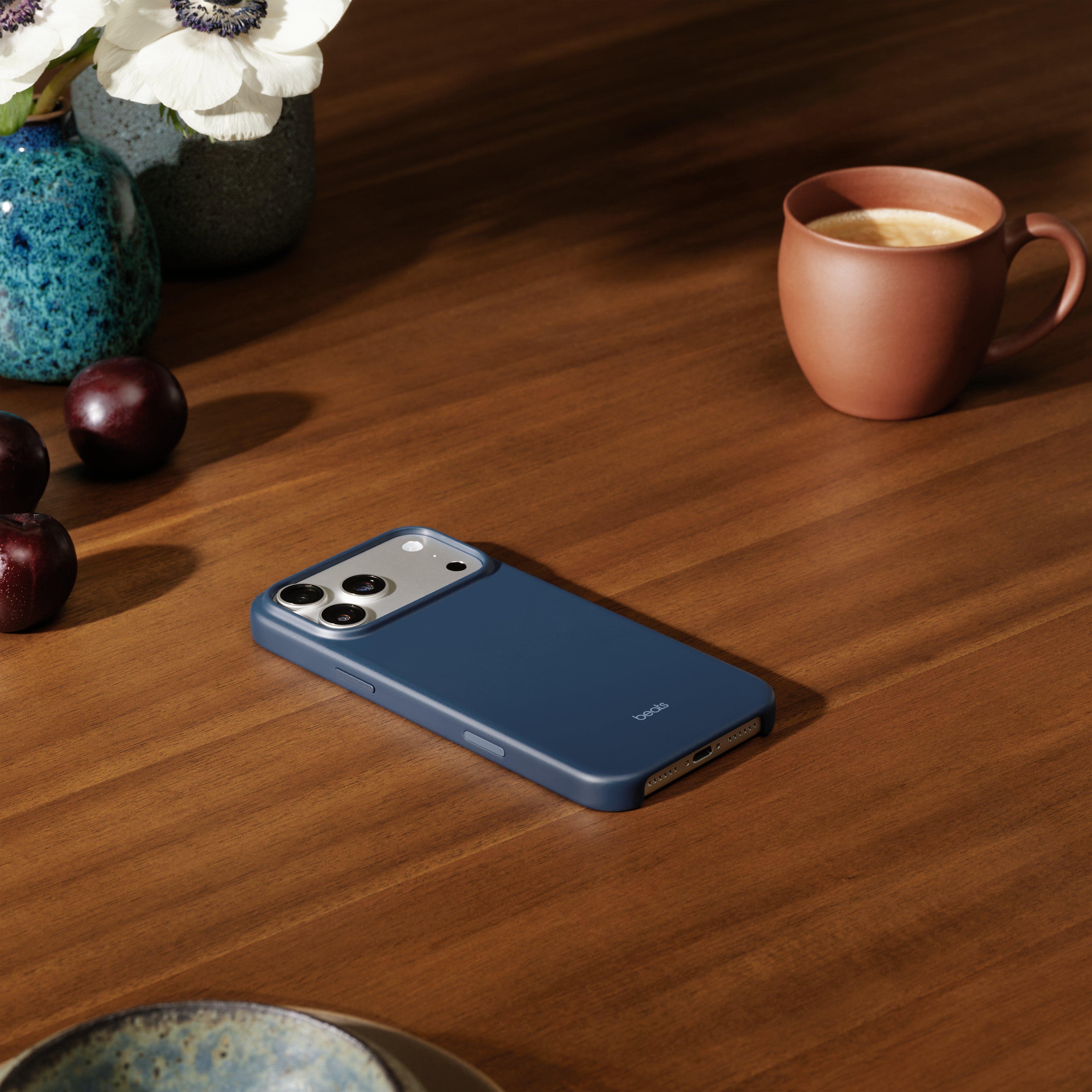 Un étui de téléphone bleu sur une table en bois avec une tasse de café, des fleurs et des prunes.