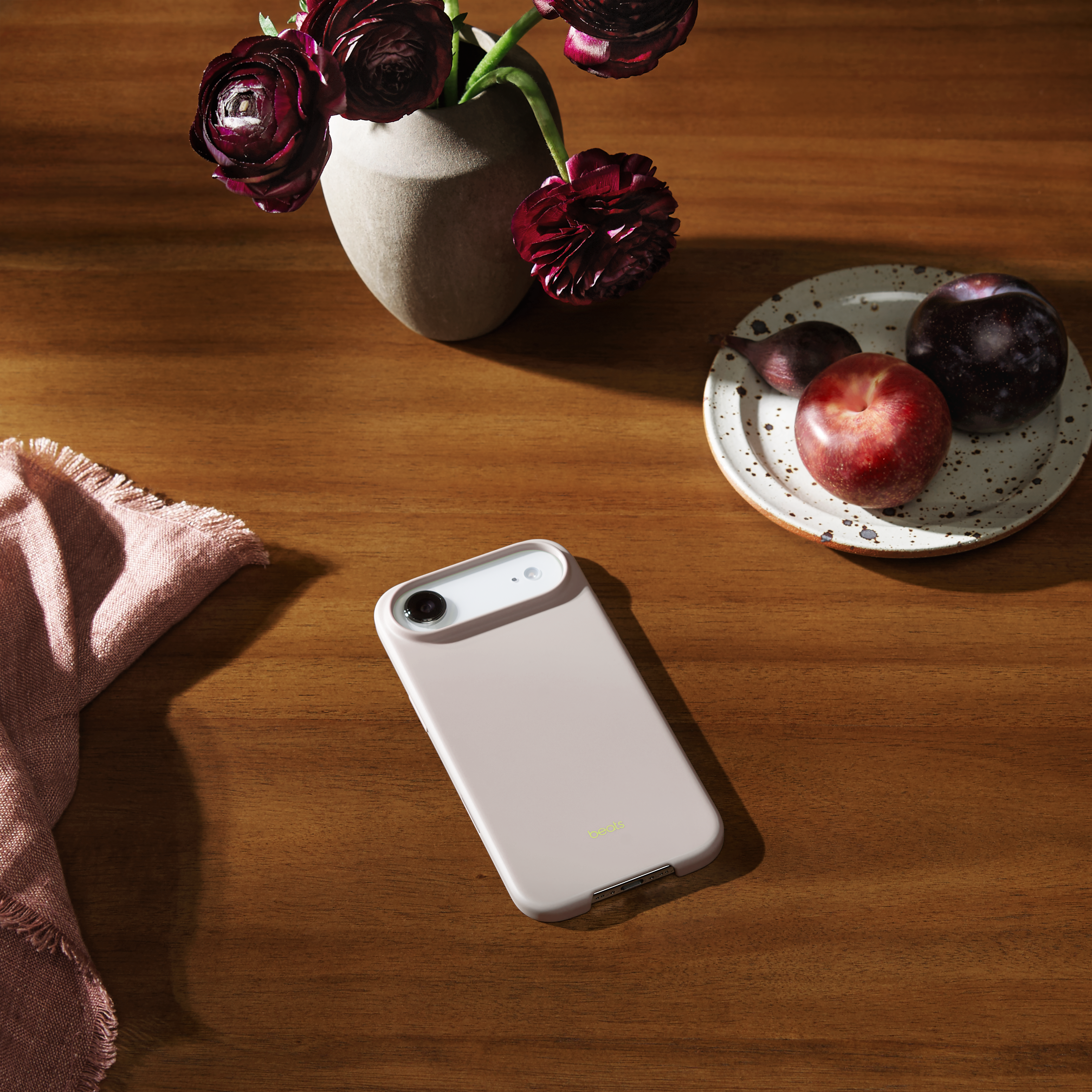 Une table en bois avec un téléphone, des fleurs dans un vase, une assiette de prunes et une serviette rose.