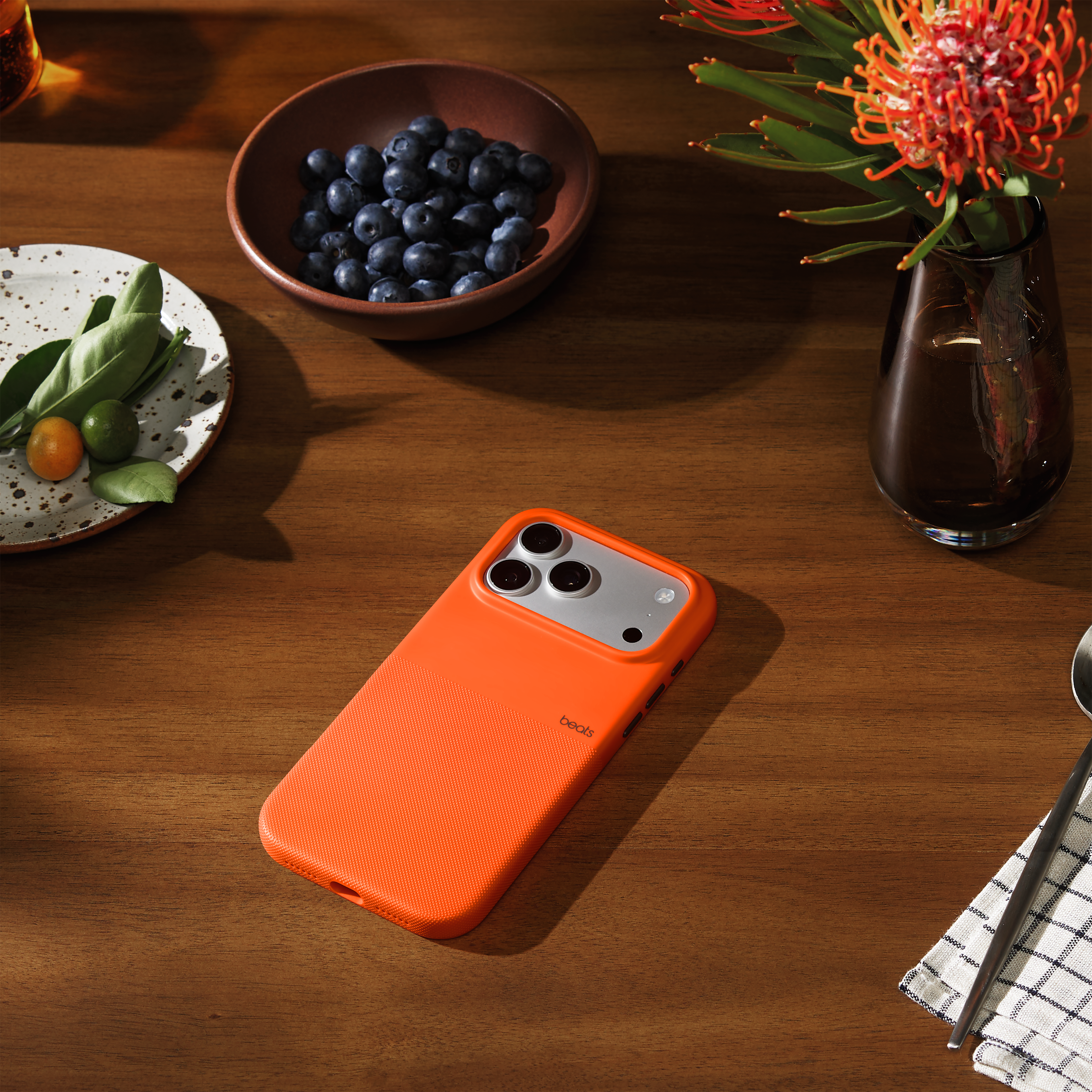 Coque de téléphone orange, myrtilles, kumquats sur une assiette, composition florale et une serviette avec des baguettes sur une table en bois.