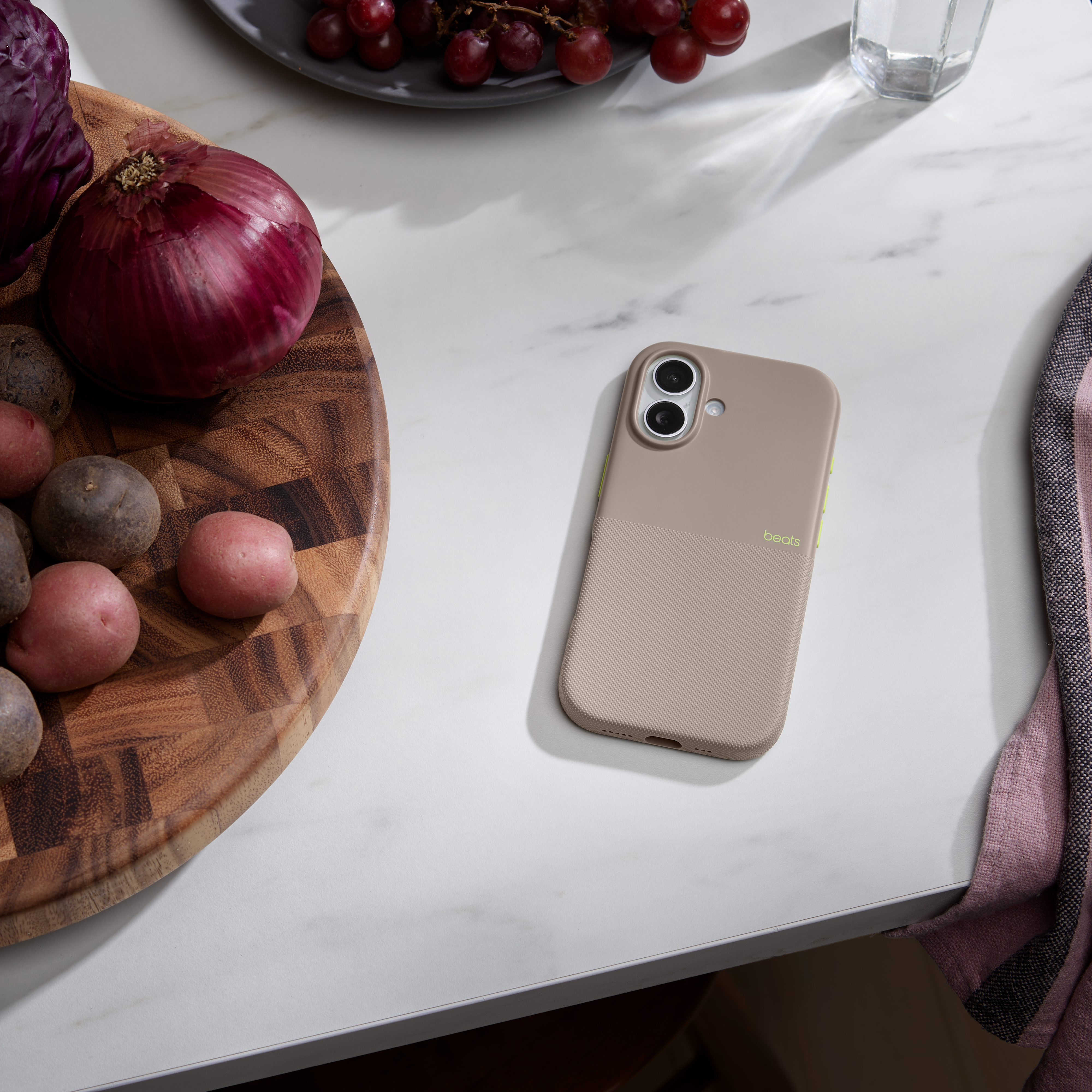 Un étui de téléphone est posé sur une table en marbre blanc. Il y a des légumes comme des oignons rouges et des pommes de terre, ainsi que des raisins et un verre.