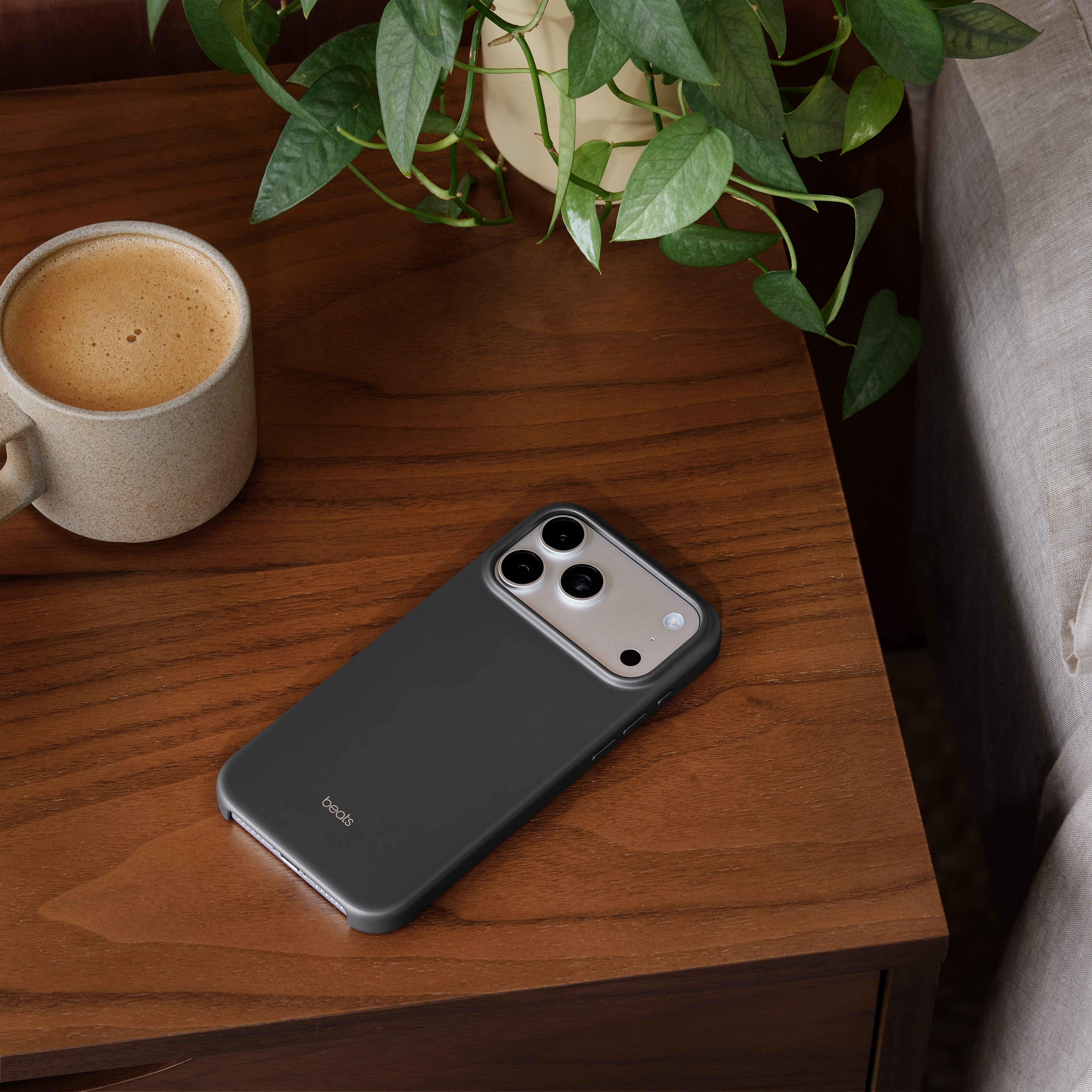 Un téléphone dans un étui noir avec une tasse de café et une plante sur une table en bois. Un canapé à droite.