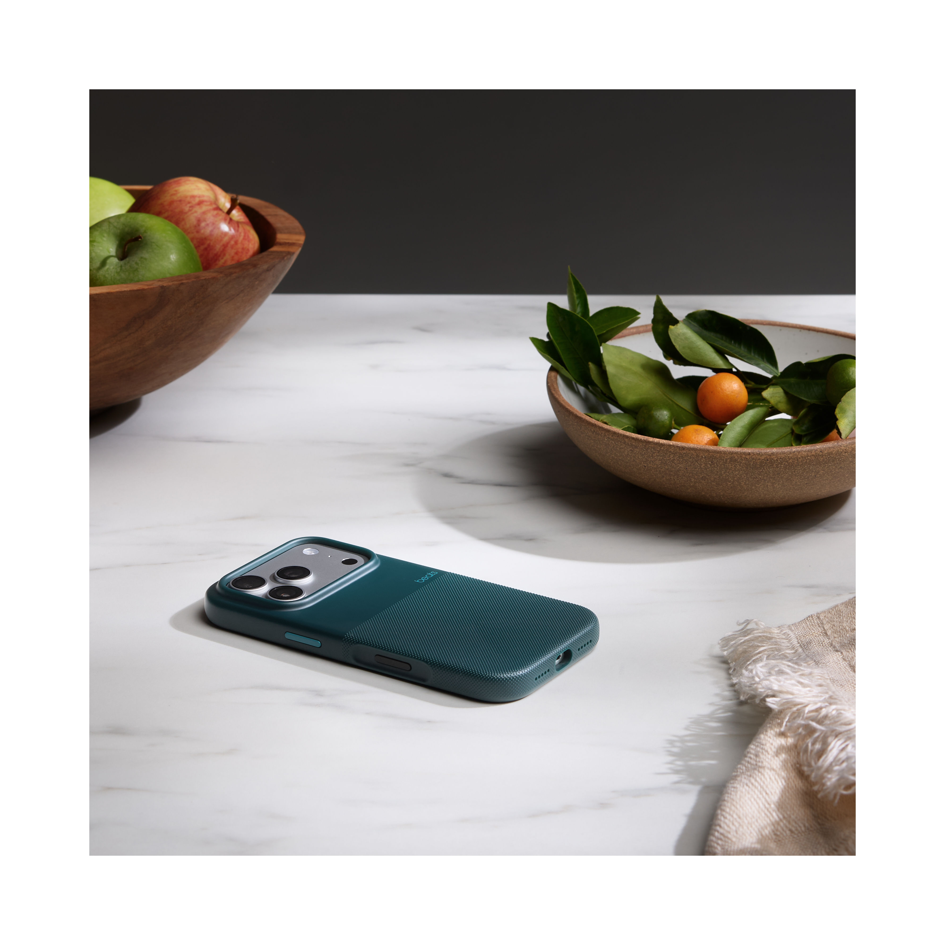 Un étui de téléphone sarcelle sur une surface en marbre. Des bols en bois avec des fruits et des feuilles en arrière-plan. Un tissu blanc à droite.