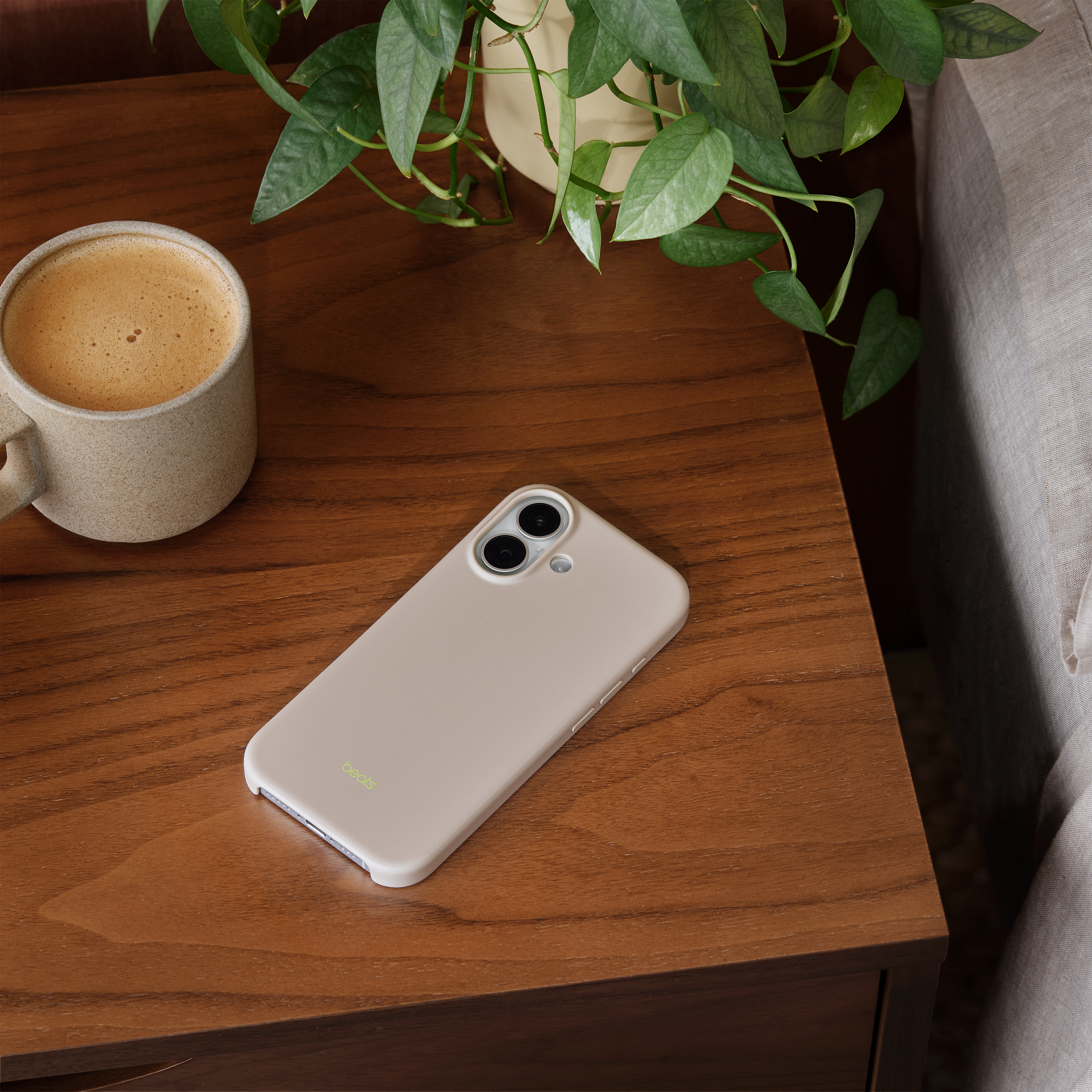 Un étui de téléphone est posé sur une table en bois avec une tasse de café et une plante. Étui et tasse beiges.