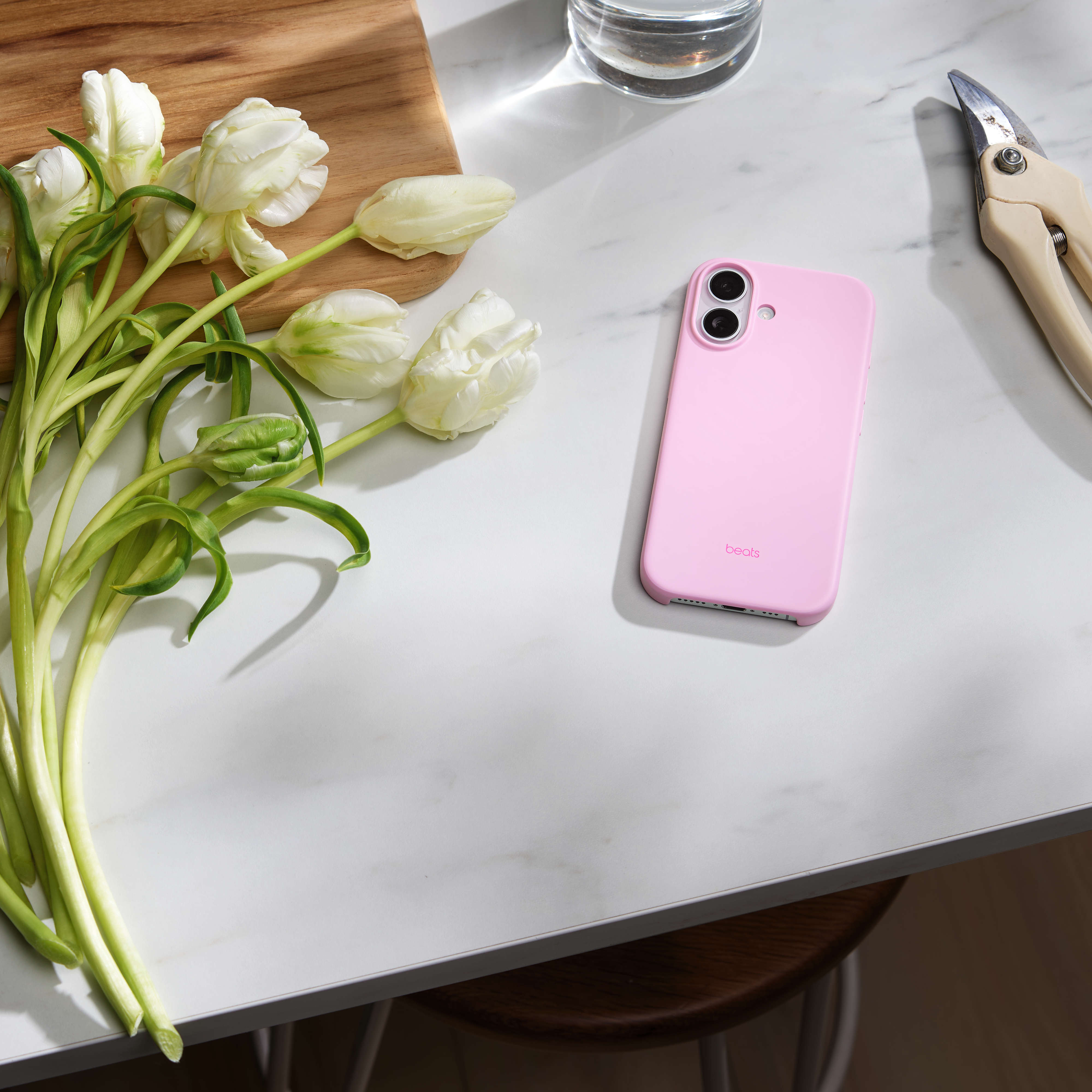 Draufsicht. Weiße Tulpen, rosa Telefon, Schere und Holzbrett auf weißer Marmoroberfläche, mit Holzstuhl im Hintergrund.