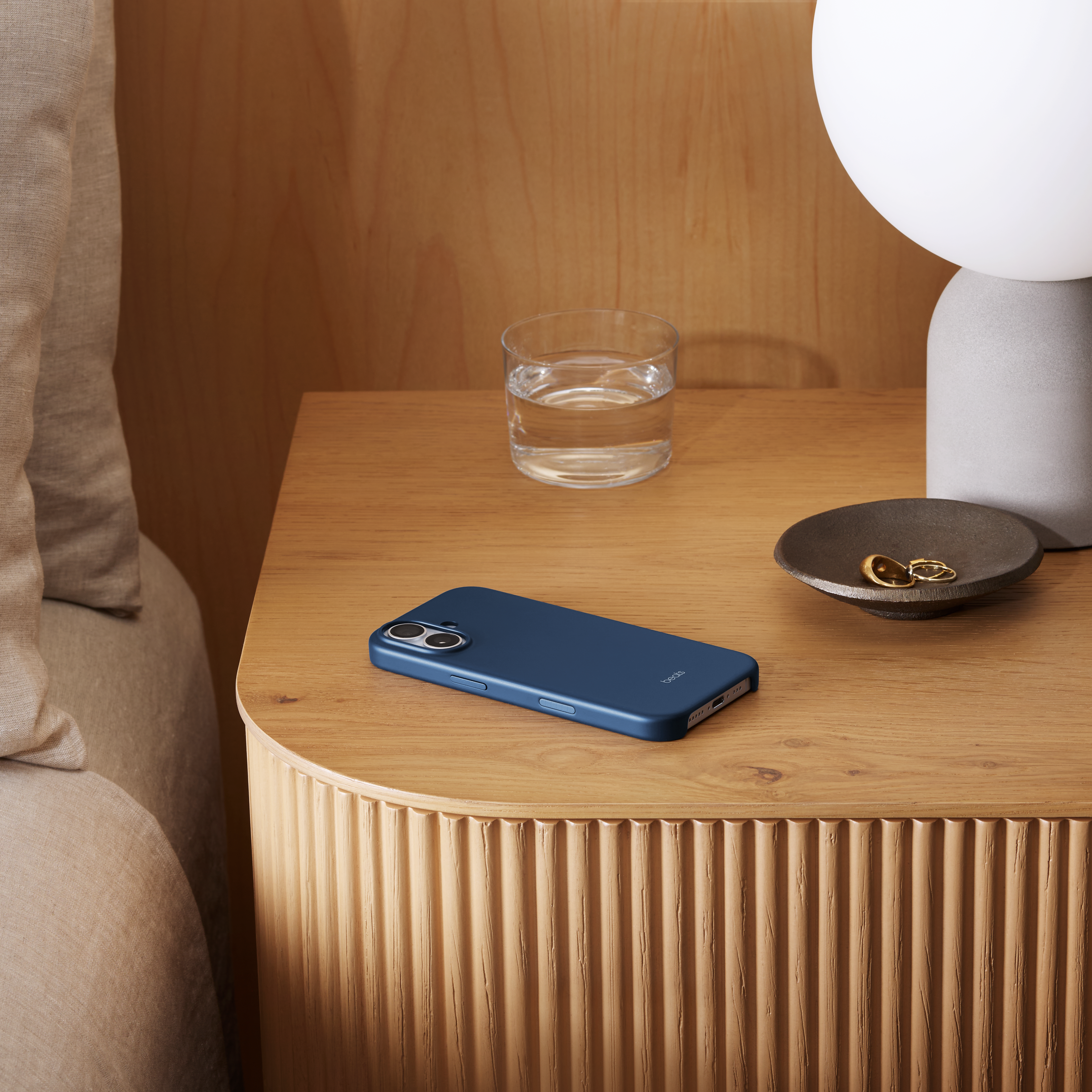 Un étui de téléphone bleu, un verre d'eau, des bijoux et une lampe sur une table de chevet en bois.