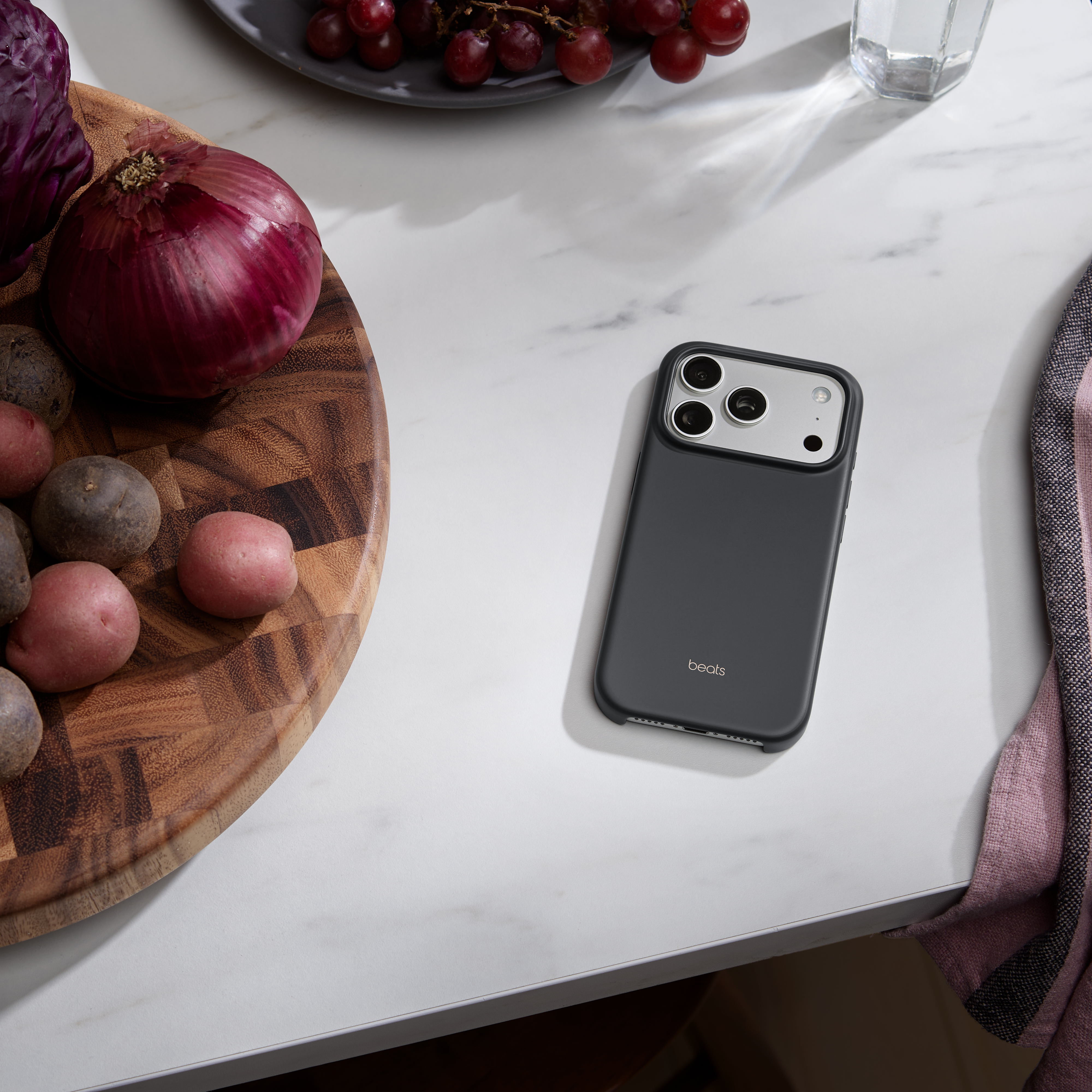 Un iPhone avec un étui Beats, oignon rouge, raisins, pommes de terre et un verre sur une surface en marbre blanc.