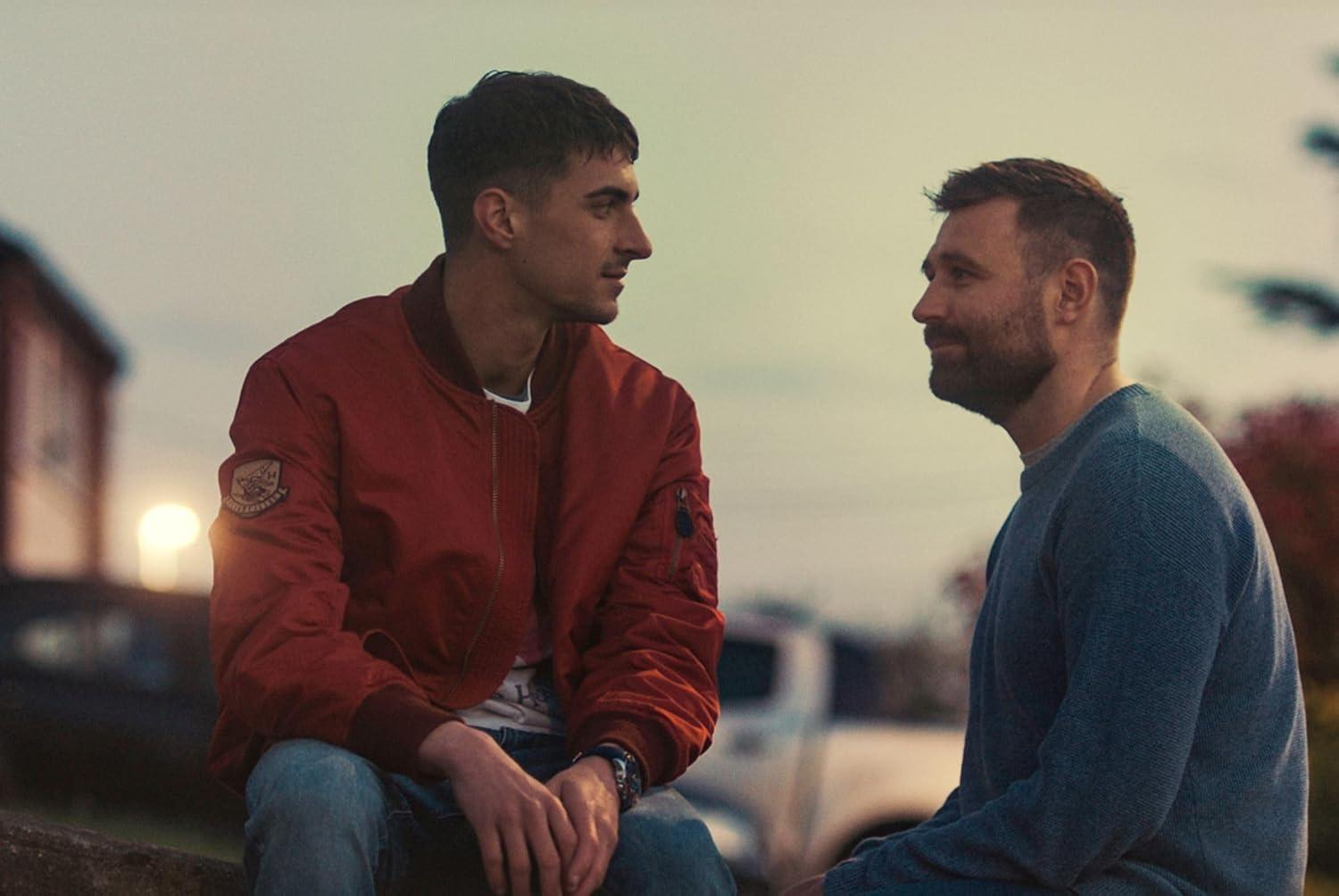 Zwei Männer sitzen sich gegenüber. Einer trägt eine rote Jacke, der andere einen blauen Pullover. Sonnenuntergang im Hintergrund.