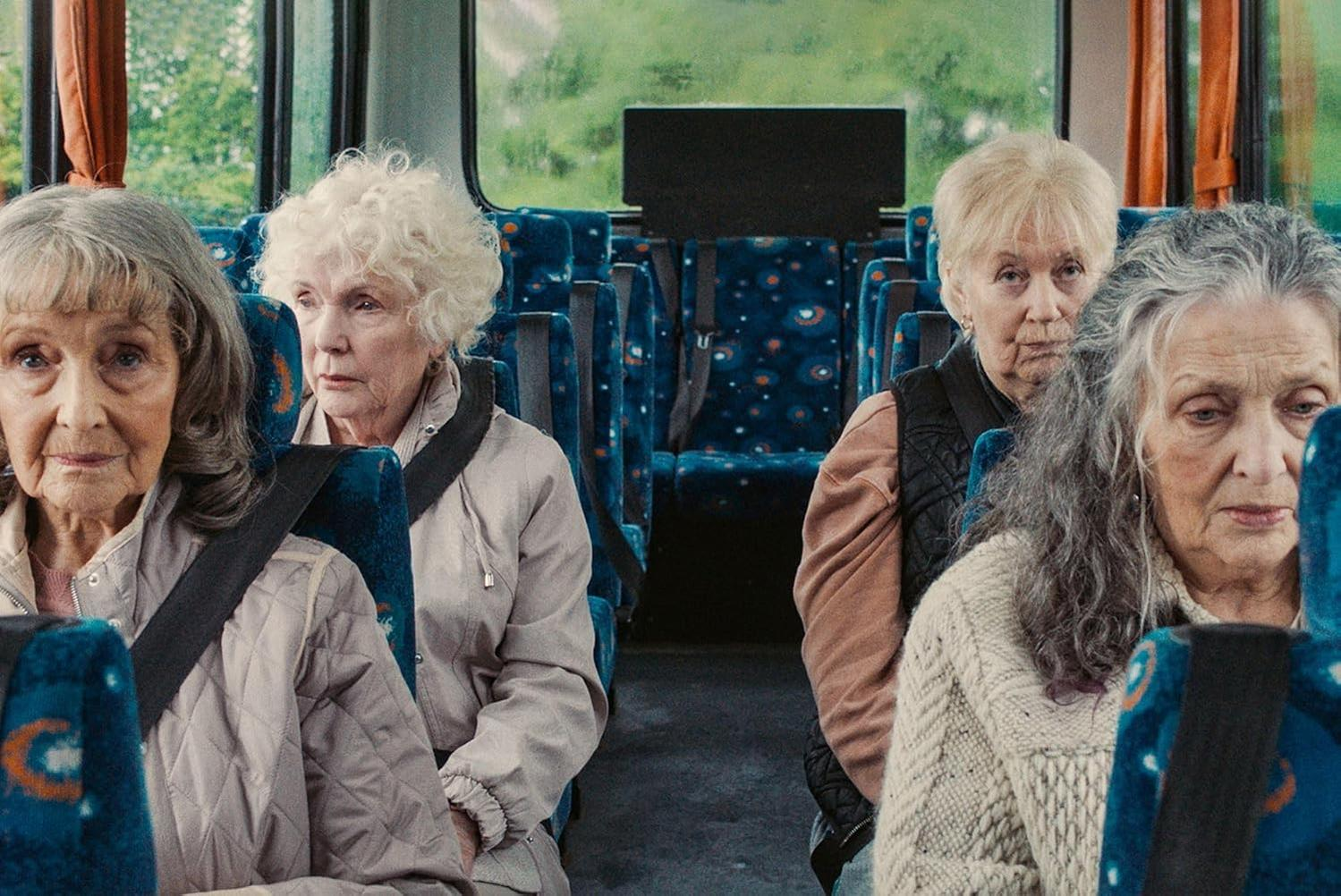 Vier ältere Frauen sitzen in einem Bus. Sie tragen Jacken. Die Sitze sind blau mit weißen Mustern. Draußen ist grüne Landschaft zu sehen.