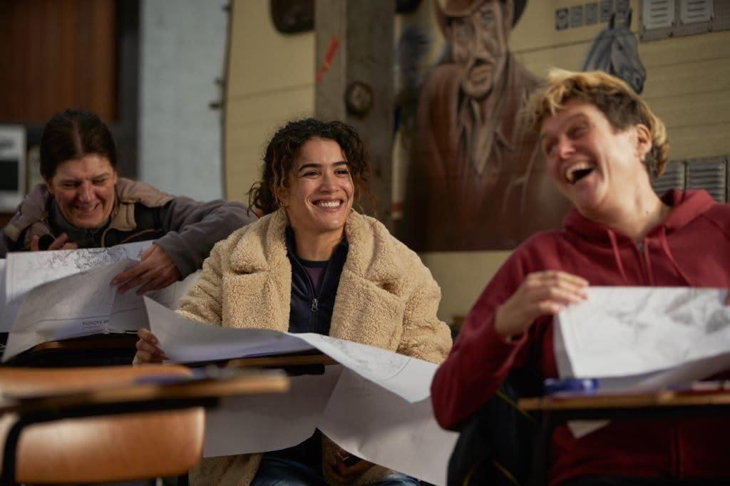 Drei Personen lachen, während sie Papiere betrachten. Einer trägt einen roten Kapuzenpullover, ein anderer einen cremefarbenen Mantel. Der Hintergrund hat ein Gemälde.
