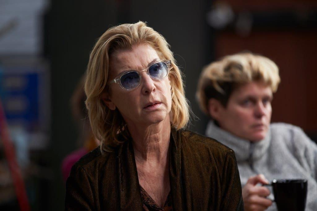 Eine Frau mit Sonnenbrille und blonden Haaren blickt in die Kamera. Eine andere Frau ist im Hintergrund verschwommen und hält eine Tasse.