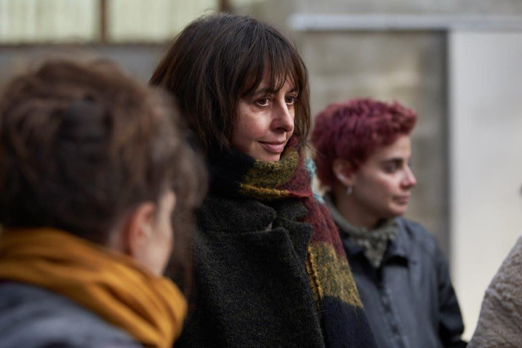 Eine Frau mit dunklem Haar und einem bunten Schal blickt zur Seite. Zwei weitere Personen sind im verschwommenen Vordergrund teilweise zu sehen.