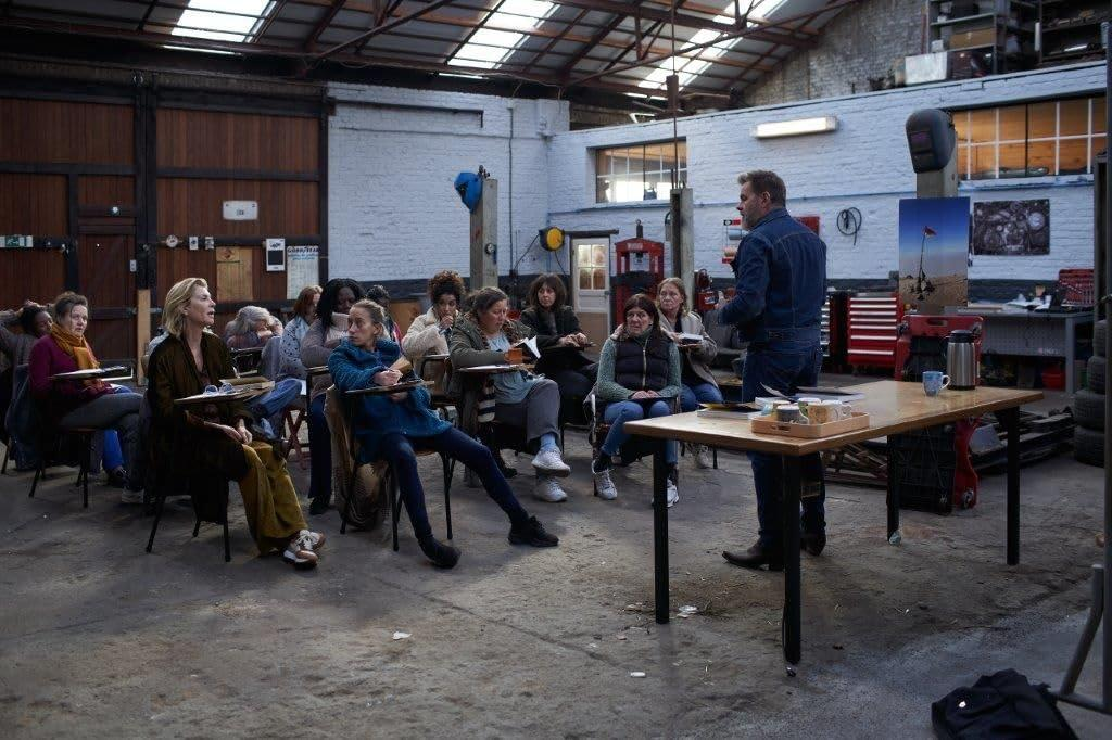 Eine Gruppe von Leuten in einer Werkstatt hört einem Mann zu. Die Leute sitzen und machen Notizen. Auf einem Tisch stehen Tassen.