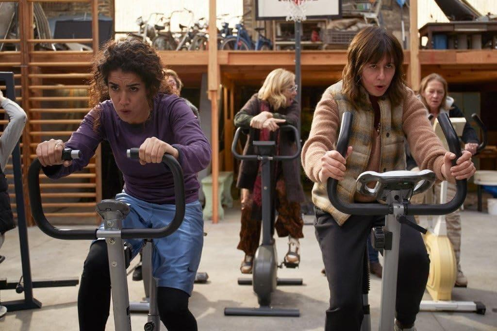 Drei Frauen auf Heimtrainern in einem Fitnessstudio. Eine Frau trägt Lila, eine andere Braun, eine weitere steht hinter ihnen.