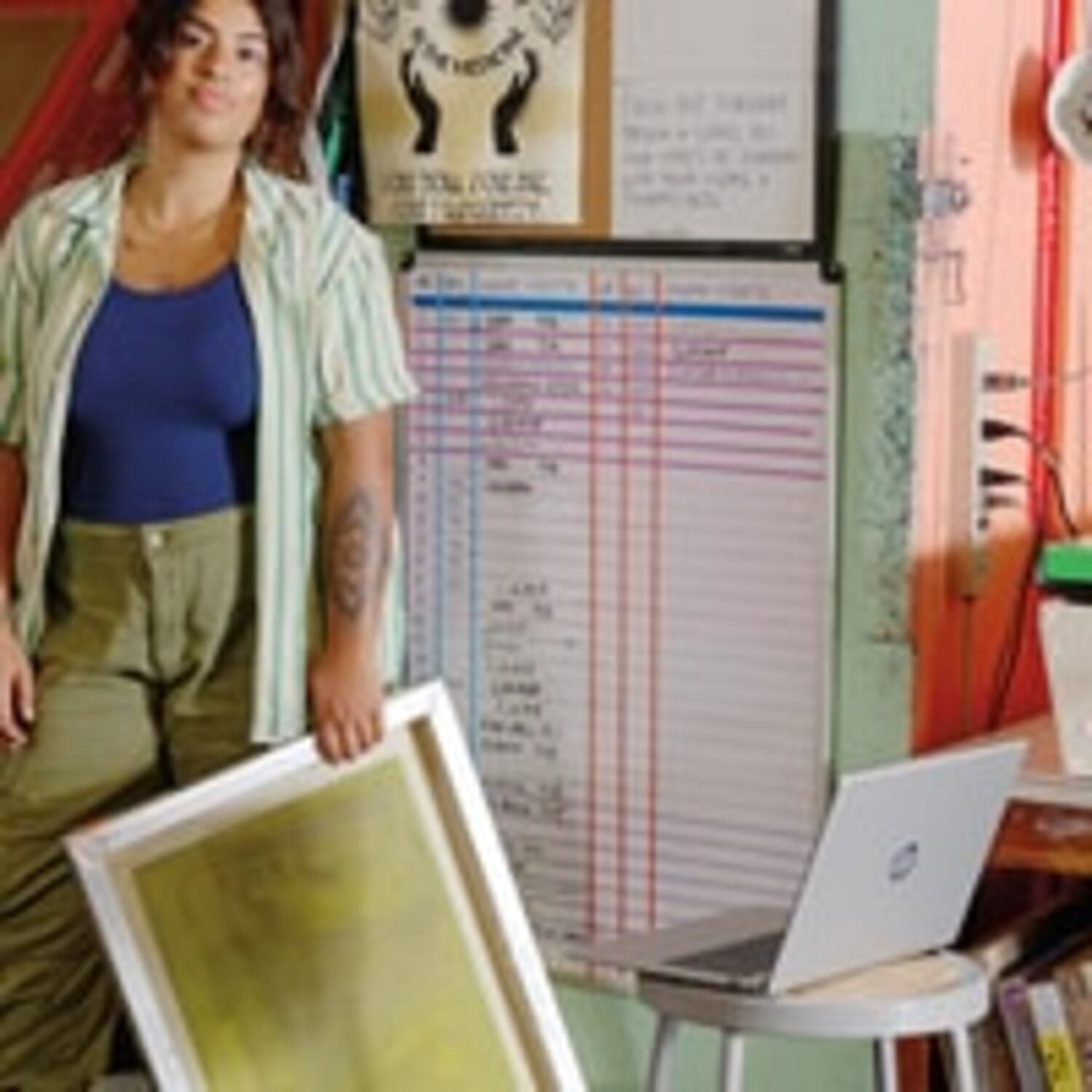 Une femme tient un écran, debout près d'un tableau blanc, d'un ordinateur portable et de livres. Elle porte un haut bleu, une chemise verte et un pantalon. Tatouage visible.