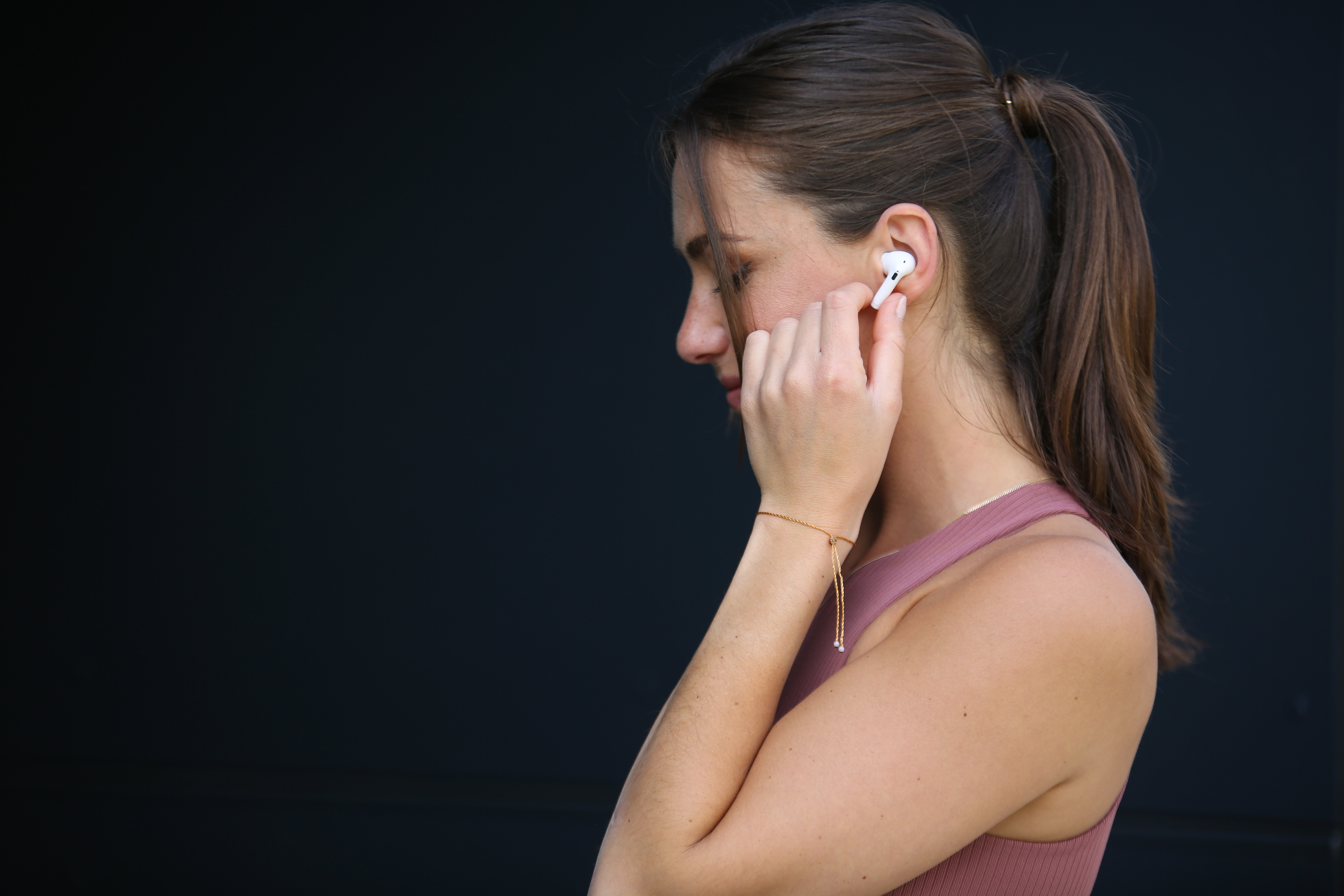 Kobieta ze słuchawką, różowym topem, kucykiem i złotą bransoletką. Ciemne tło.