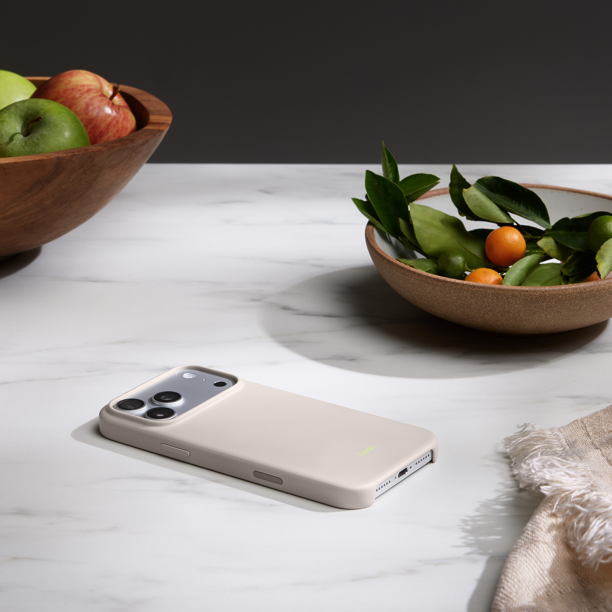 Beżowe etui na telefon leży na marmurowej powierzchni. Miski z owocami i liśćmi w tle. Beżowa szmatka po prawej stronie.