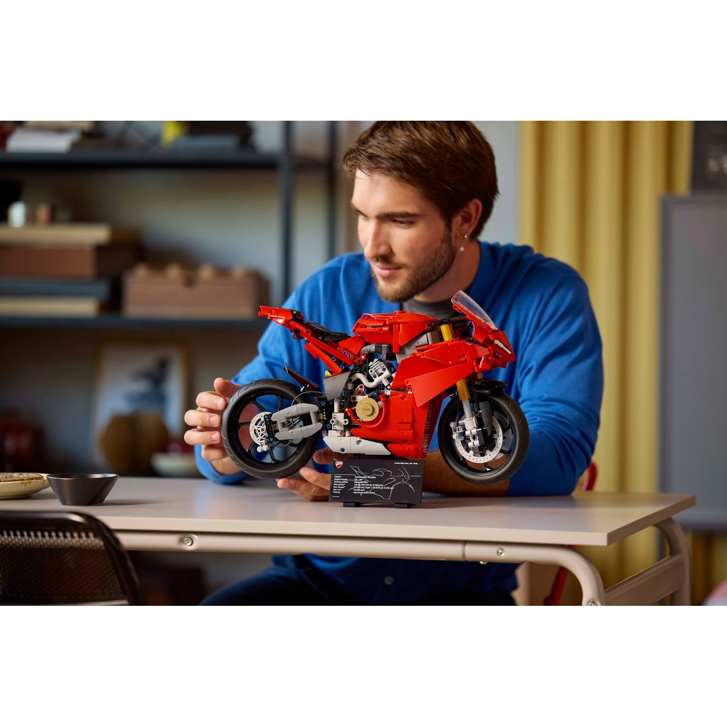 Un uomo tiene in mano un modello di moto Lego rosso su un tavolo. Sorride, indossa una camicia blu. Sfondo: scaffali e un muro.