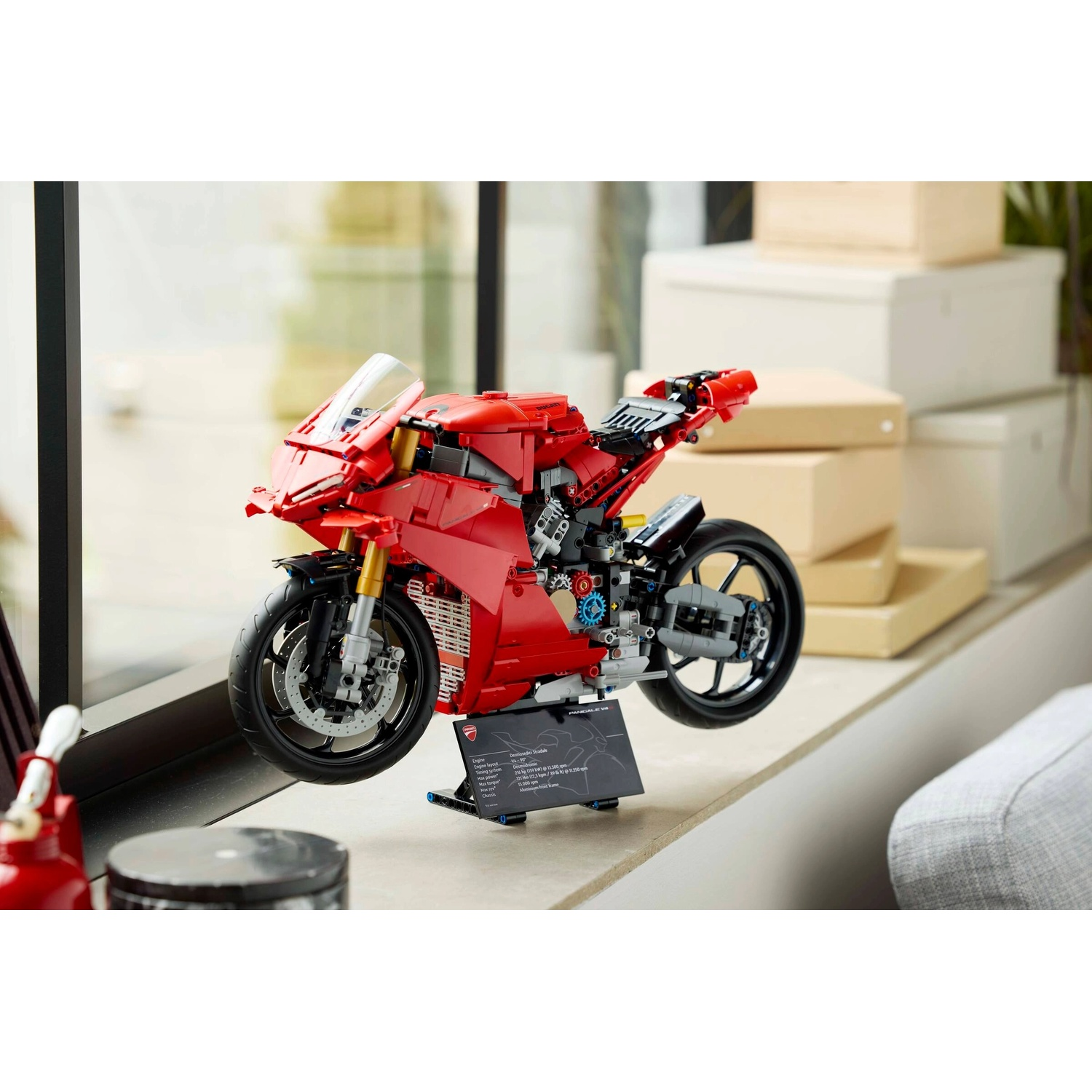 Un modello di moto Ducati Lego rosso è esposto su un davanzale. Scatole di cartone dietro. Una targa informativa nera è davanti.