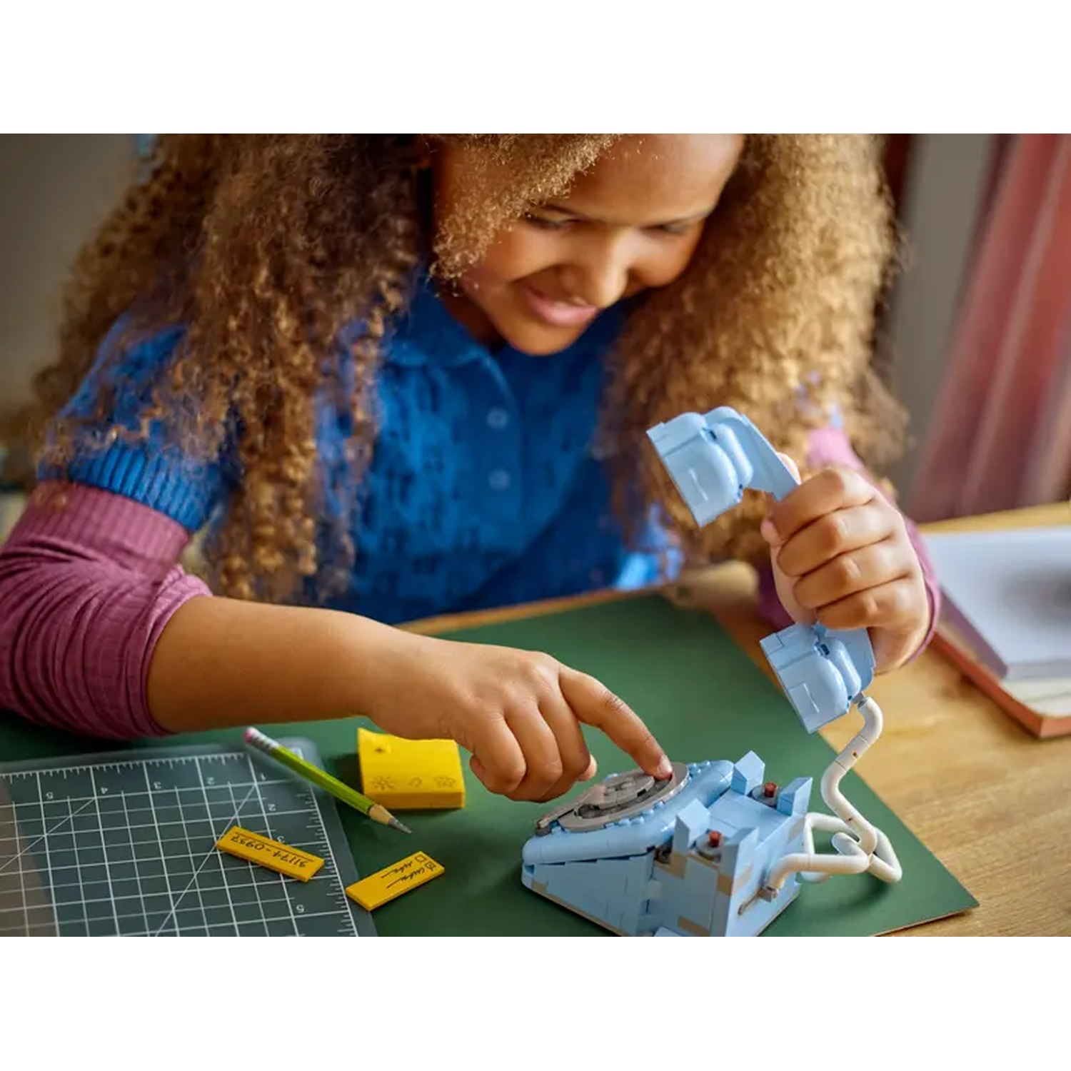Una ragazza sta assemblando un telefono Lego blu. Gli accessori includono una matita, post-it gialli e un tappetino da taglio verde.