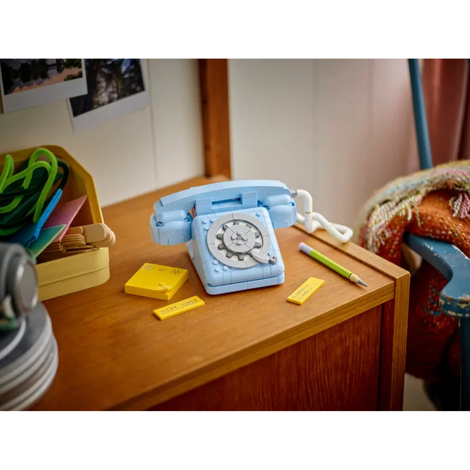 Un telefono LEGO blu è su una scrivania di legno con post-it gialli e una matita. Si vede una scatola con materiali per l'artigianato.