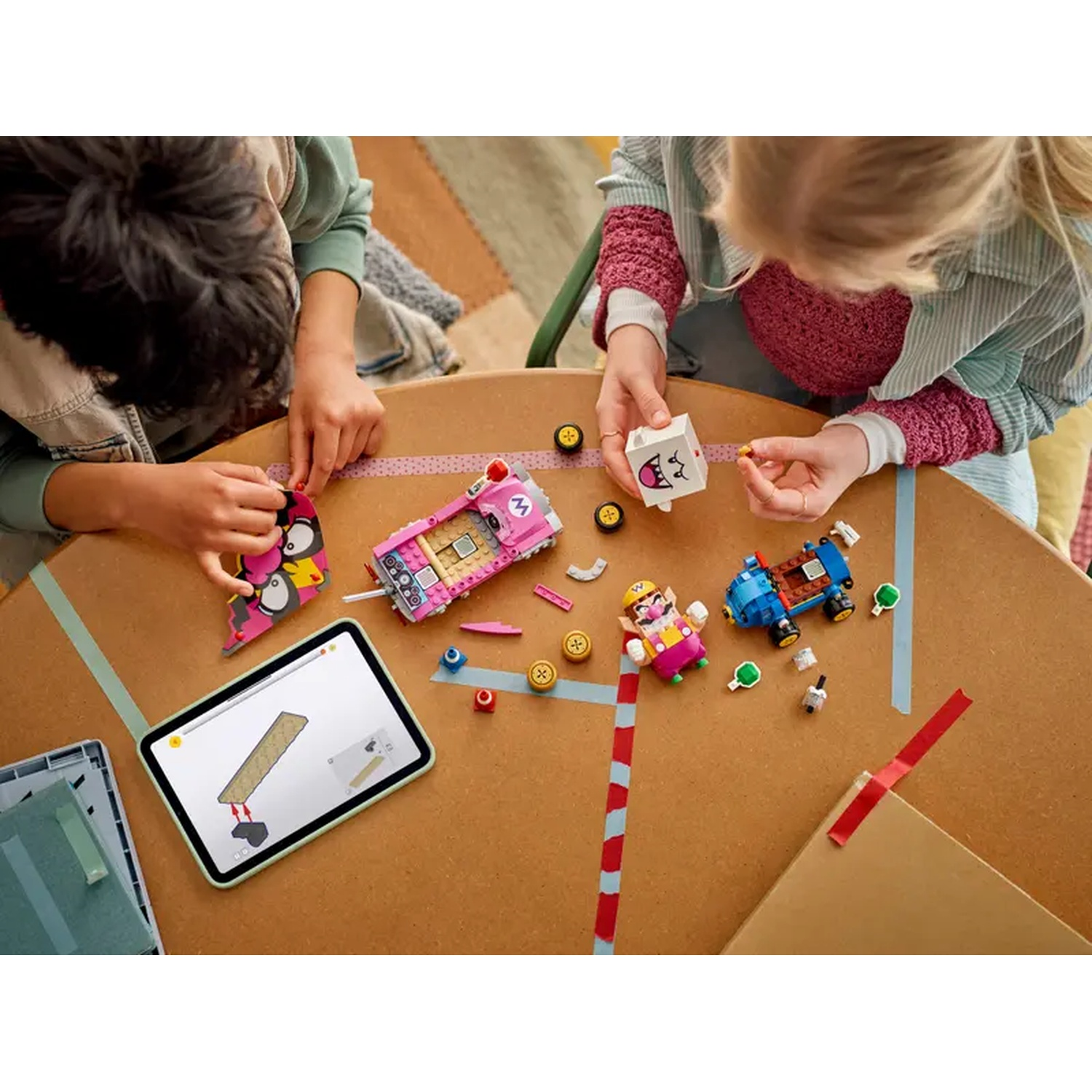 Bambini che costruiscono con giocattoli Lego su un tavolo marrone, tra cui un'auto rosa, una blu e un tablet con istruzioni.
