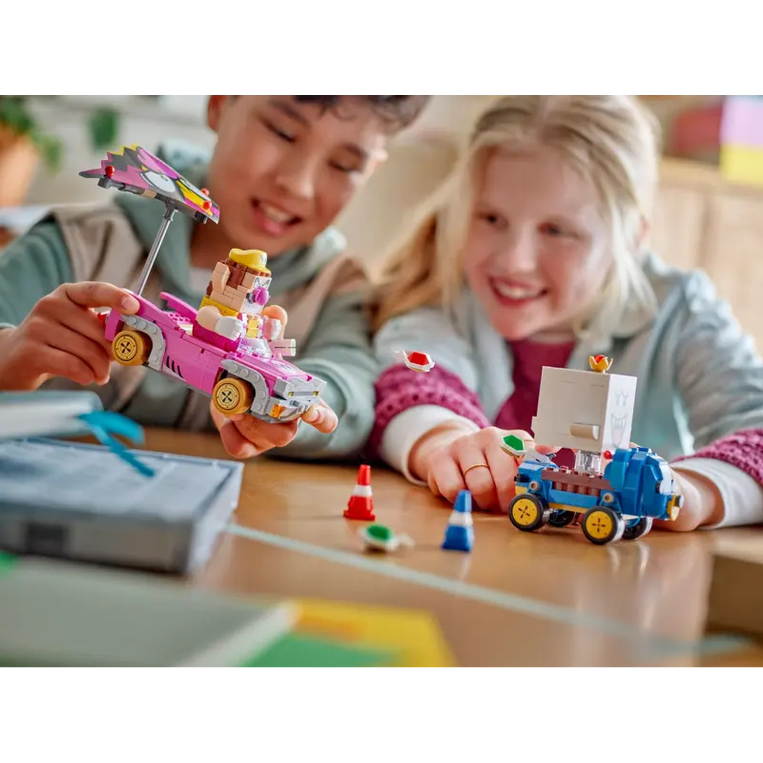 Due bambini giocano con le auto LEGO. Un'auto rosa con un ombrello e un camion blu con una scatola bianca sono su un tavolo.