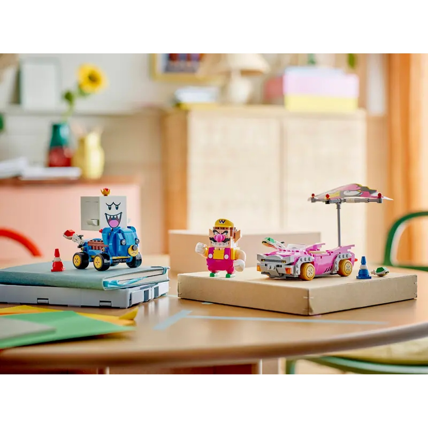 Personaggi Lego di Wario e un Boo che guidano veicoli su un tavolo, con mobili e un girasole sullo sfondo.