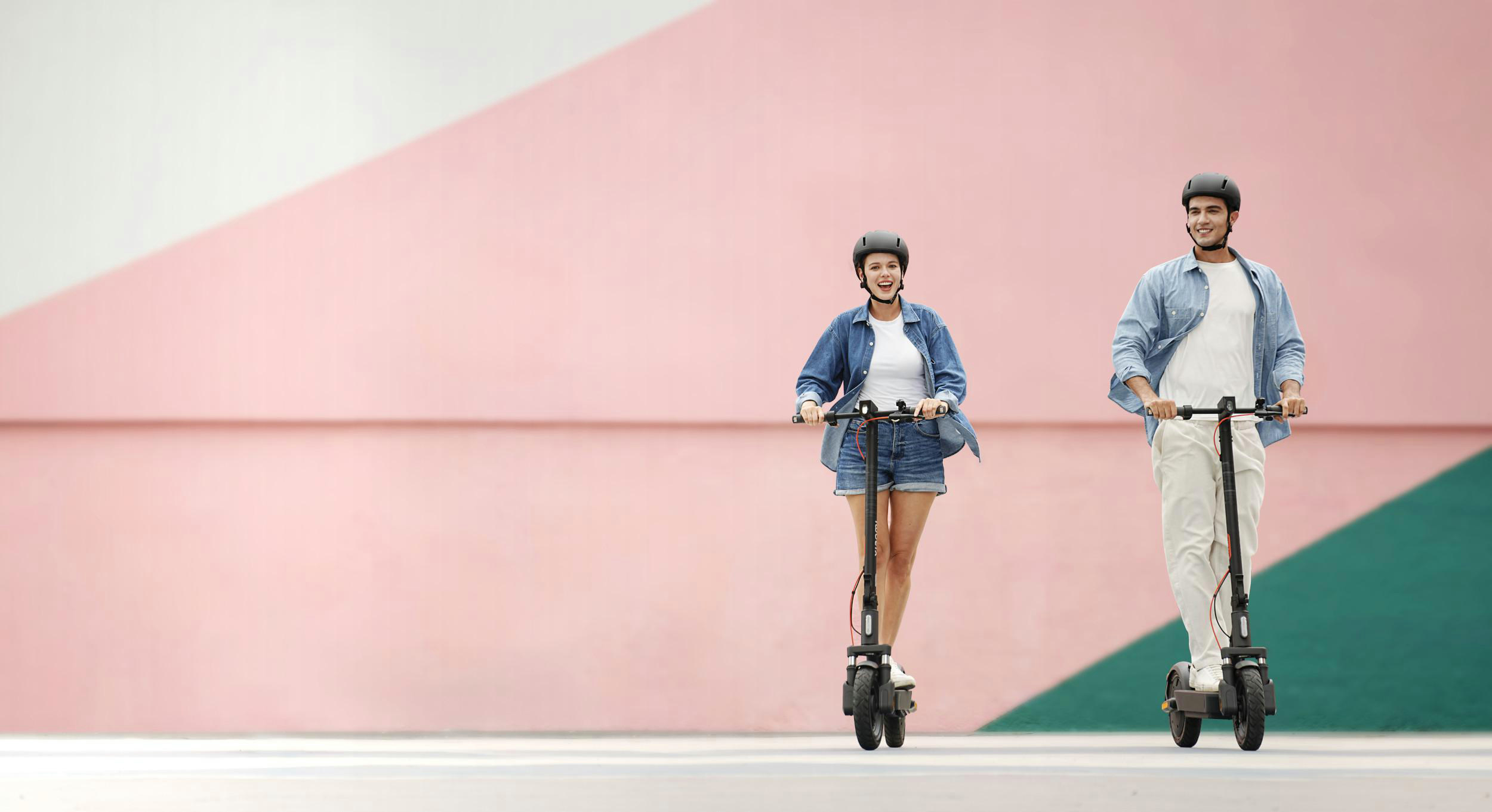 Zwei Personen auf Elektrorollern, mit Helmen, fahren vor einem rosa und weißen Hintergrund.