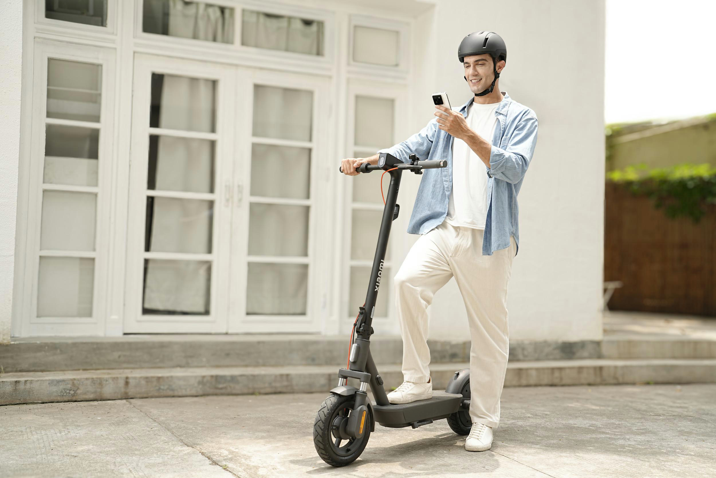 Mann auf Roller, Helm, Telefon. Weiße Hose, blaues Hemd. Hintergrund: weiße Türen, Stufen und Gebäude.