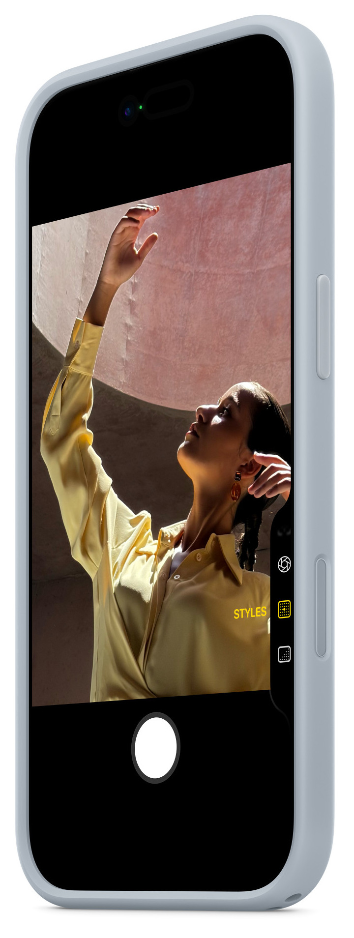 Kobieta w żółtej koszuli unosi rękę w kierunku różowego tła, widocznego przez ekran telefonu. Telefon jest jasnoniebieski.