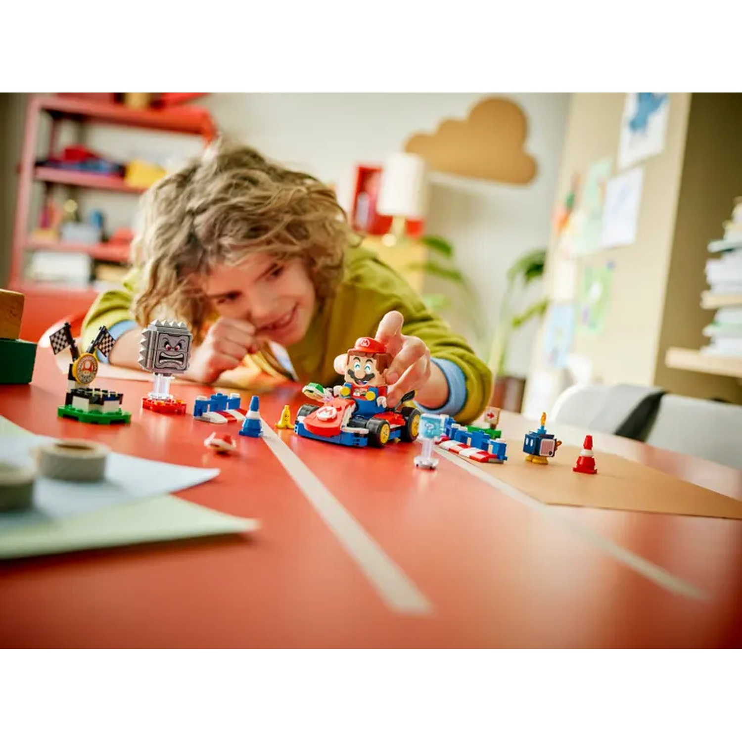 Un bambino gioca con un set Lego di Mario. Mario è in un kart rosso, altre figure Lego sono su un tavolo rosso. Il bambino sorride.