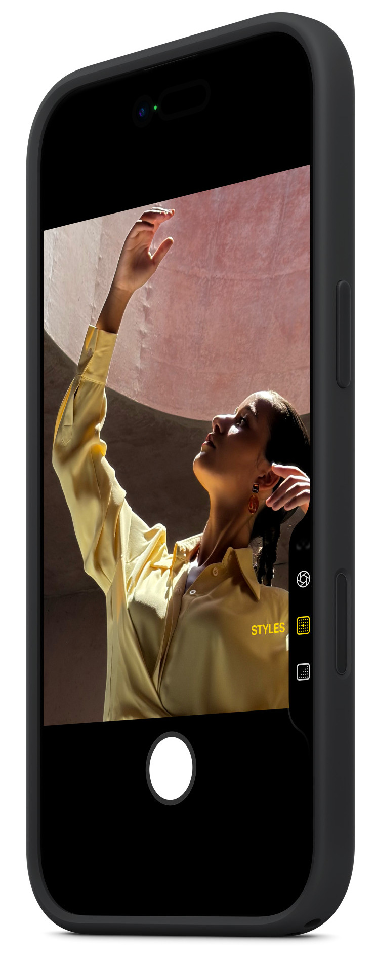 Kobieta w żółtej koszuli, wyciąga rękę. Obraz uchwycony na ekranie telefonu, z różową ścianą w tle.