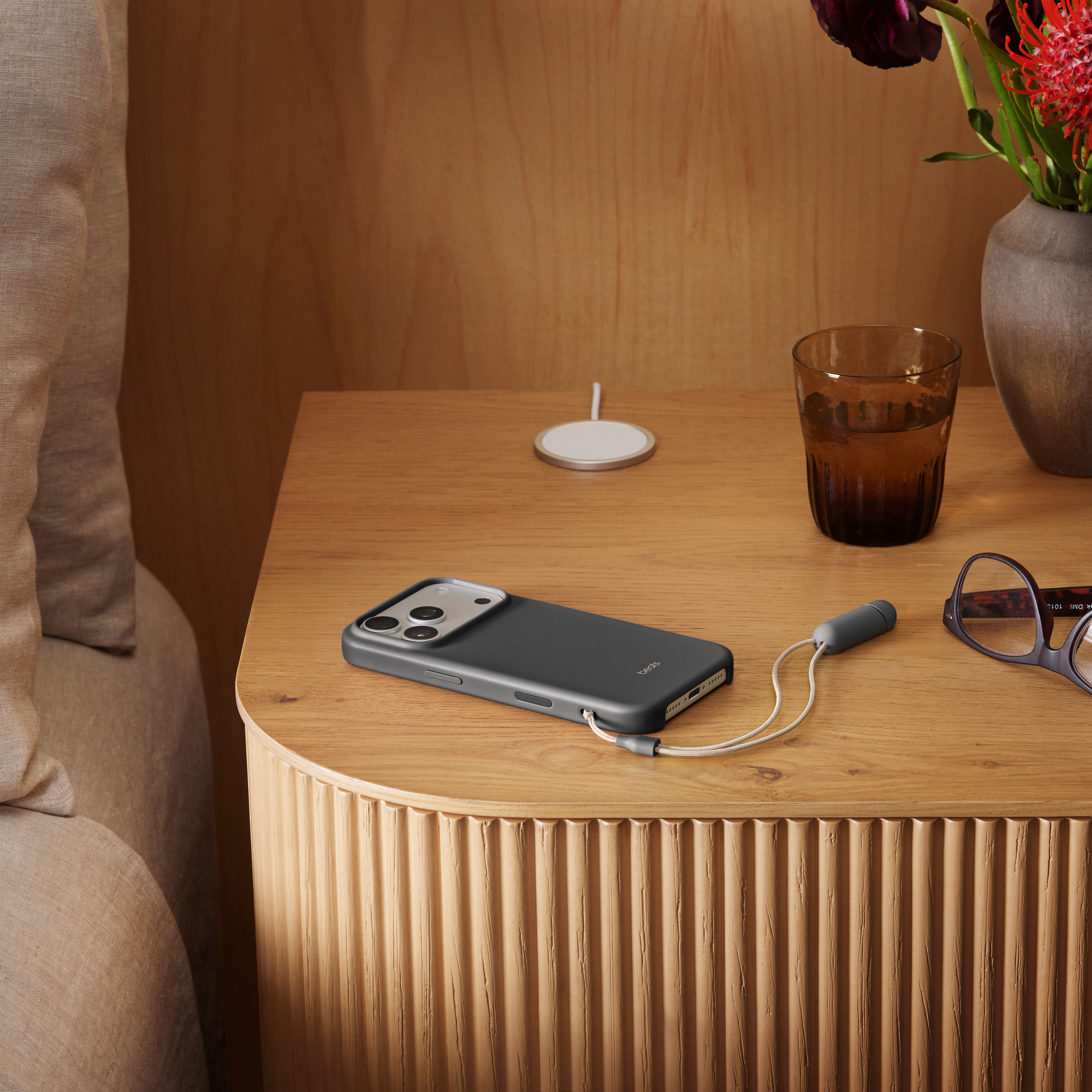 Une table de chevet avec un téléphone, un câble, un verre et des fleurs. Un lit à gauche.