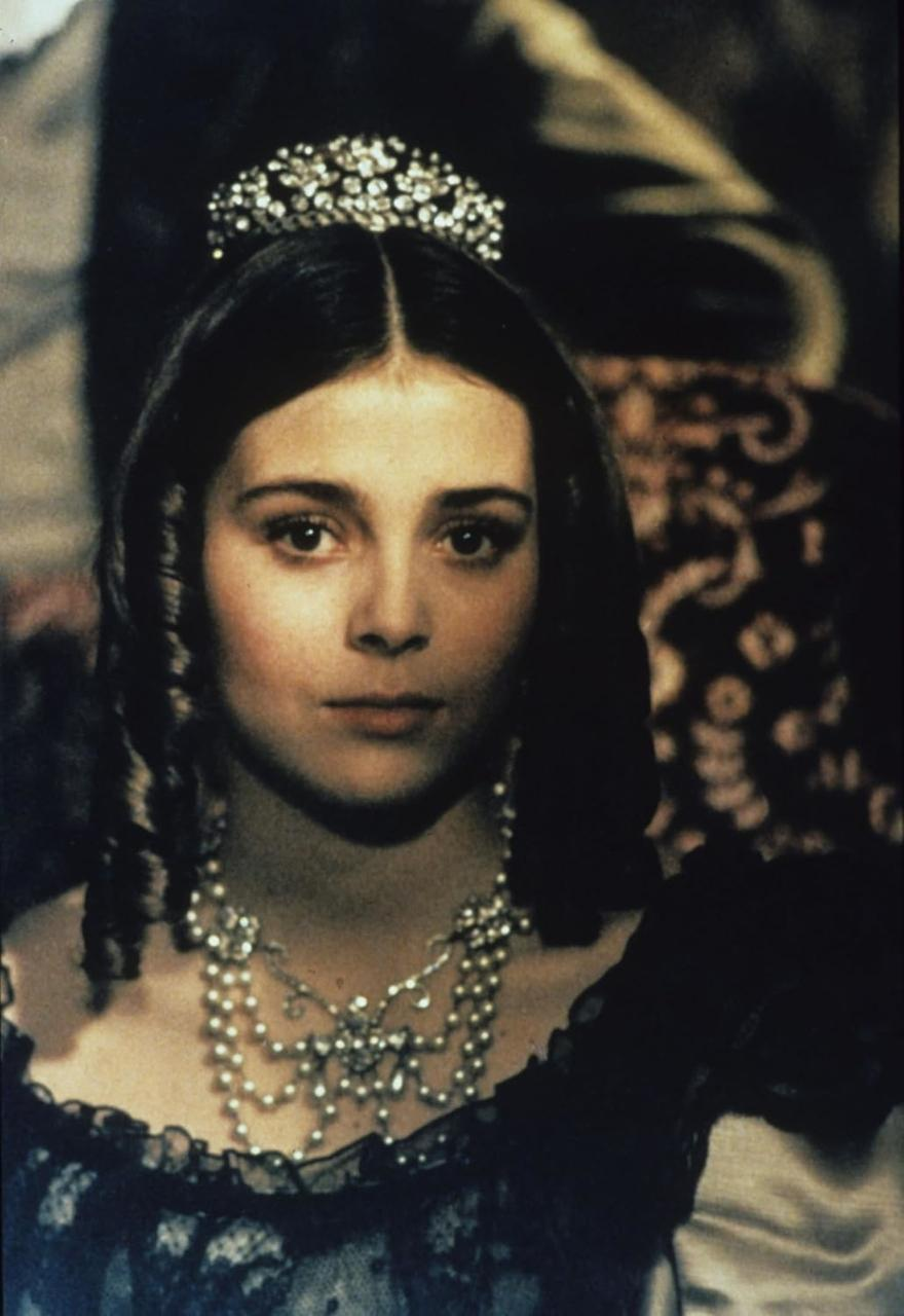 Eine Frau mit dunklem Haar, die ein juwelenbesetztes Diadem und eine Halskette trägt, blickt den Betrachter an. Sie trägt ein dunkles Kleid. Der Hintergrund ist unscharf.