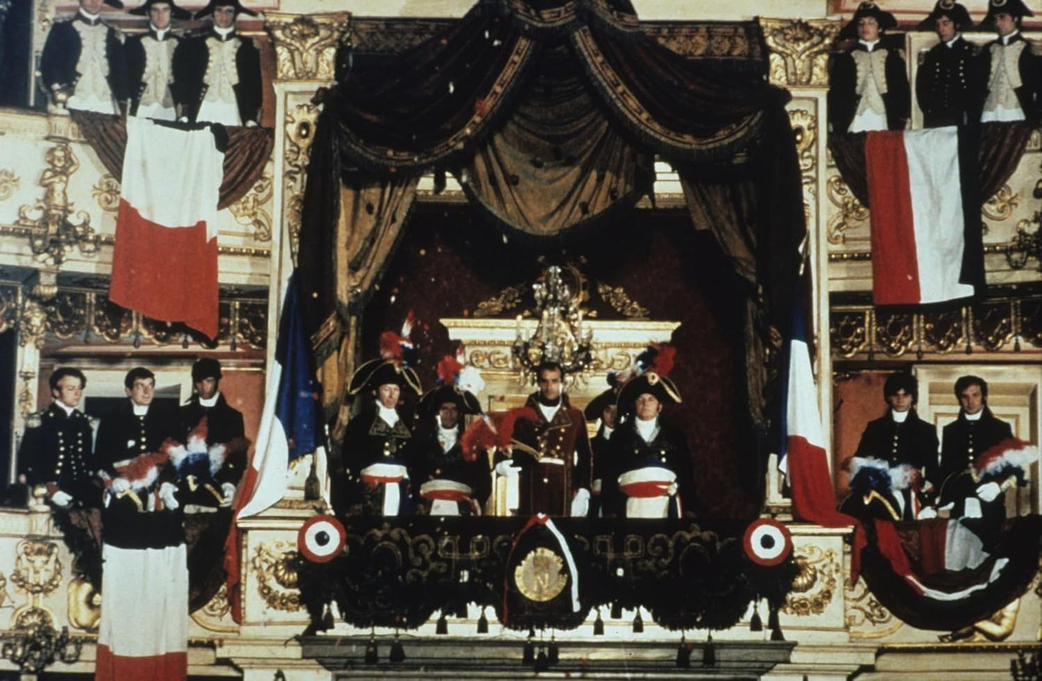 Zeremonie mit Männern in historischer Kleidung, Flaggen und aufwendigen Verzierungen. Menschen stehen auf einem Balkon, möglicherweise eine historische Nachstellung.