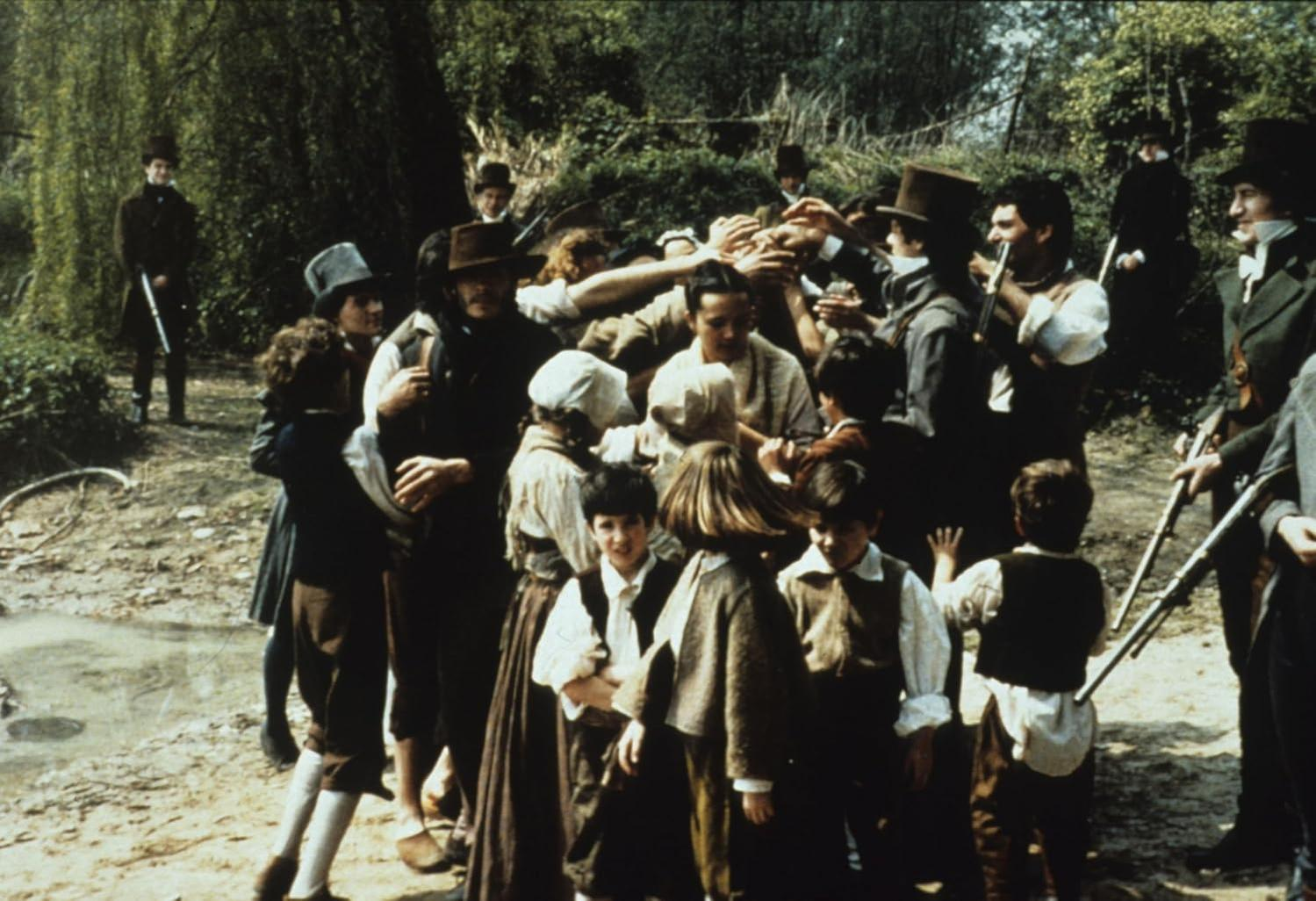 Eine Gruppe von Menschen in historischer Kleidung versammelt sich an einem Fluss. Einige halten Gewehre. Kinder sind im Vordergrund, Erwachsene im Hintergrund.