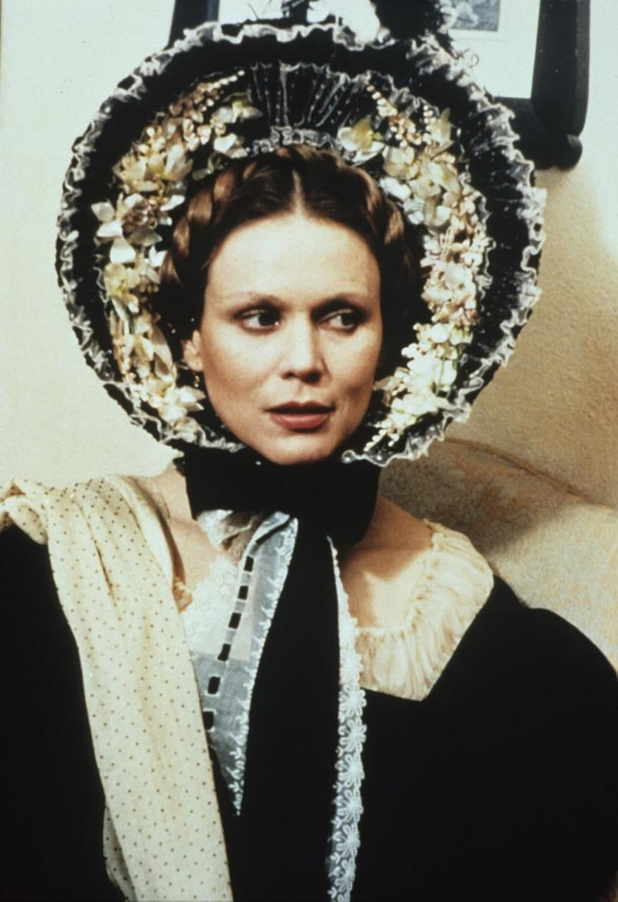 Frau mit Blumenschmuck-Hut und schwarzem Kleid mit Spitzenkragen und schwarzer Schleife. Sie schaut zur Seite.