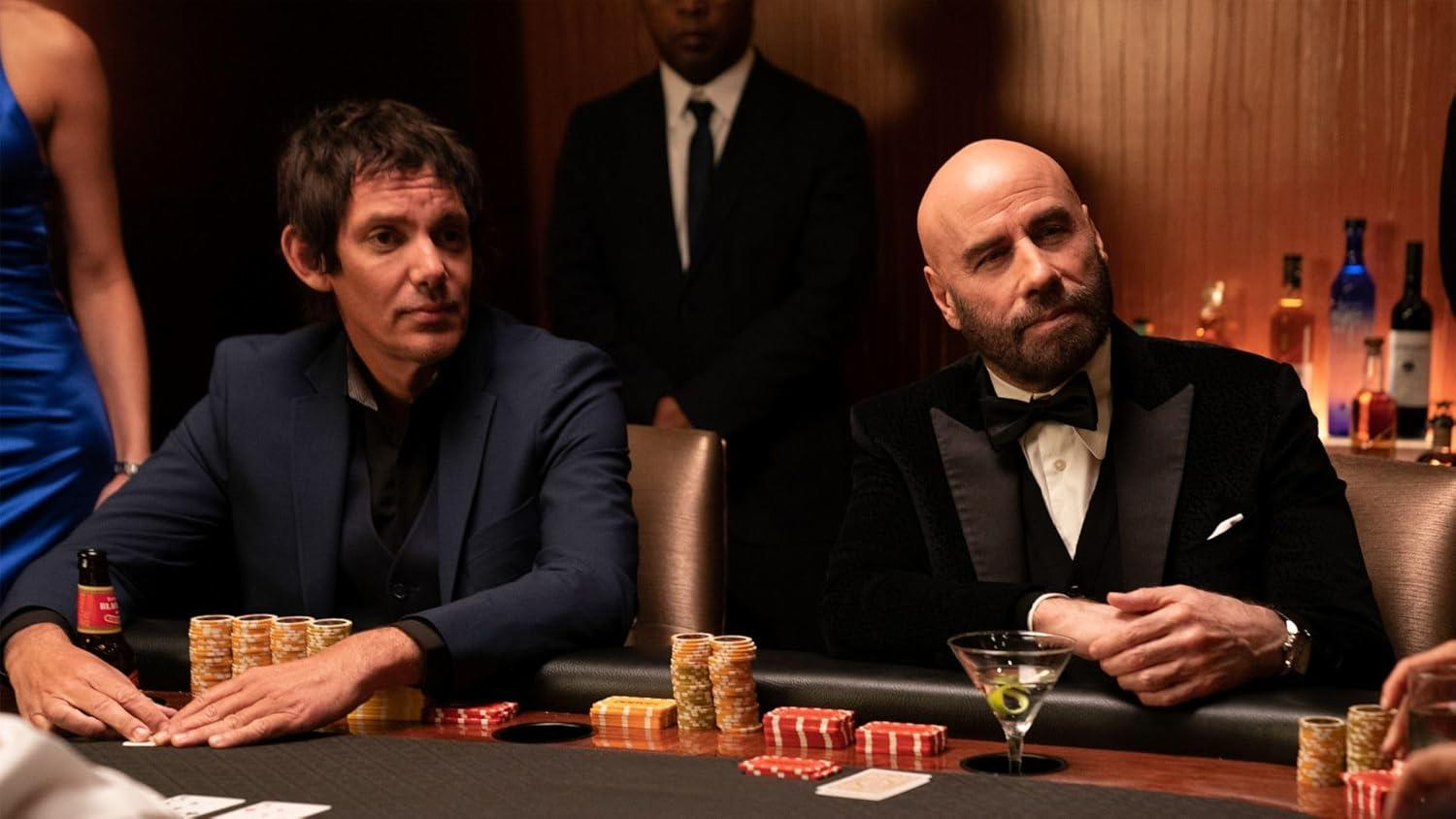 Zwei Männer an einem Pokertisch, einer im Anzug, der andere im Smoking. Chips, Karten und Getränke liegen auf dem Tisch. Ein Kellner steht hinter ihnen.