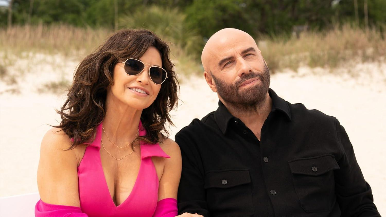Eine Frau in Pink und ein kahler Mann in Schwarz sitzen am Strand. Die Frau trägt eine Sonnenbrille. Dahinter ist ein Sandstrand.