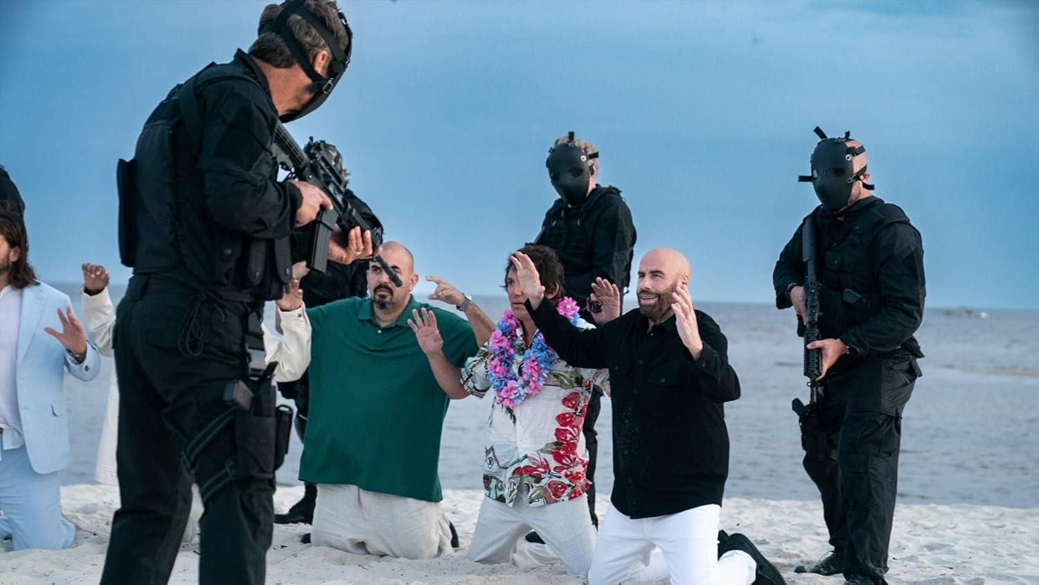 Männer mit erhobenen Händen am Strand, konfrontiert mit bewaffneten Gestalten in schwarzen Masken. Der Himmel ist blau. Die Szene erzeugt Spannung.