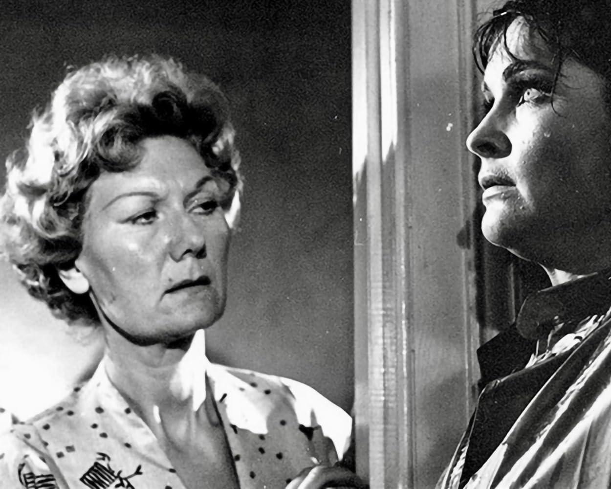 Zwei Frauen in einem Schwarzweißfilm. Eine wirkt besorgt, die andere starrt nach vorn. Beide befinden sich in einem Raum mit einer Tür.