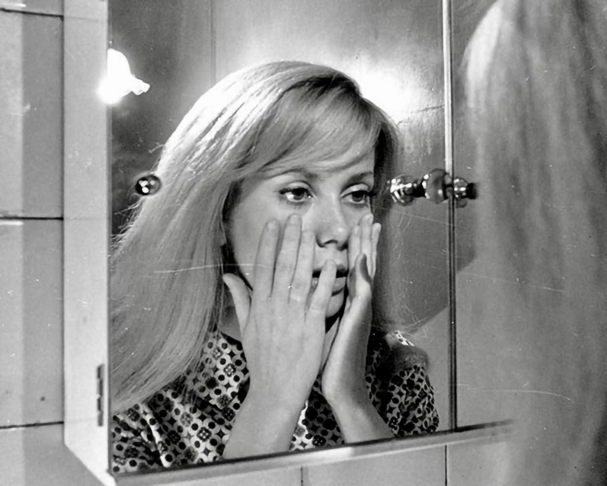 Eine Frau betrachtet sich im Spiegel, die Hände im Gesicht. Sie hat blondes Haar und trägt ein gemustertes Hemd in einem Badezimmer.