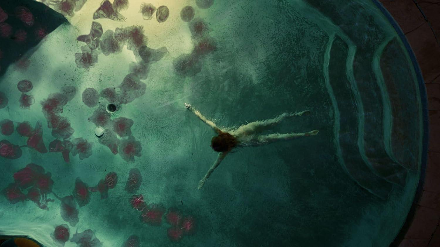 Eine Person schwimmt in einem Pool mit grünem Wasser. Rötliche Formen schweben nahe der Oberfläche. Die Poolränder sind sichtbar.
