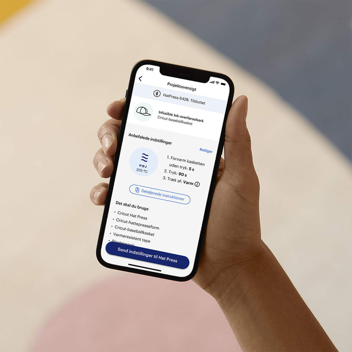 Eine Hand hält ein Smartphone, das einen Cricut Hat Press-Bildschirm mit Anweisungen anzeigt. Text und ein Baseballkappen-Symbol sind sichtbar.