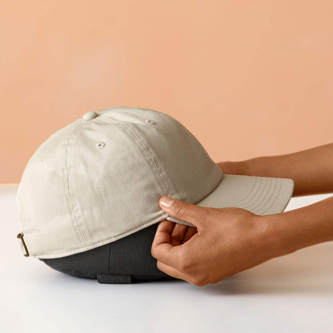 Hände halten eine beige Baseballkappe über einem dunkelgrauen Objekt vor einem pfirsichfarbenen Hintergrund auf einer weißen Oberfläche.