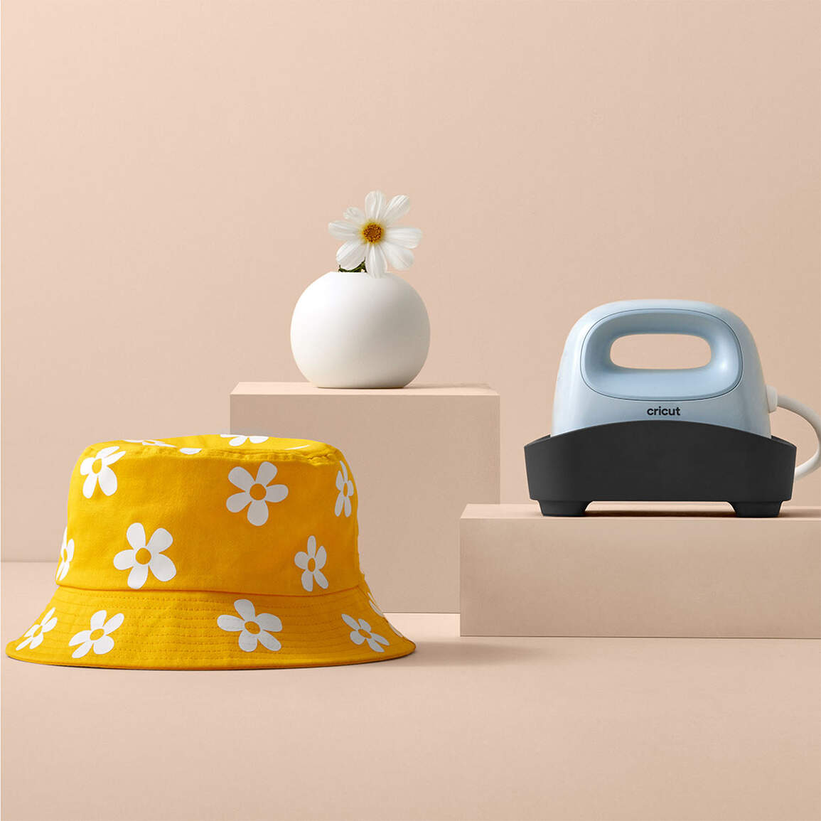 Ein gelber Fischerhut mit weißen Gänseblümchenmustern, eine weiße Vase mit einer Gänseblümchen und ein Cricut-Bügeleisen auf beigen Podesten.