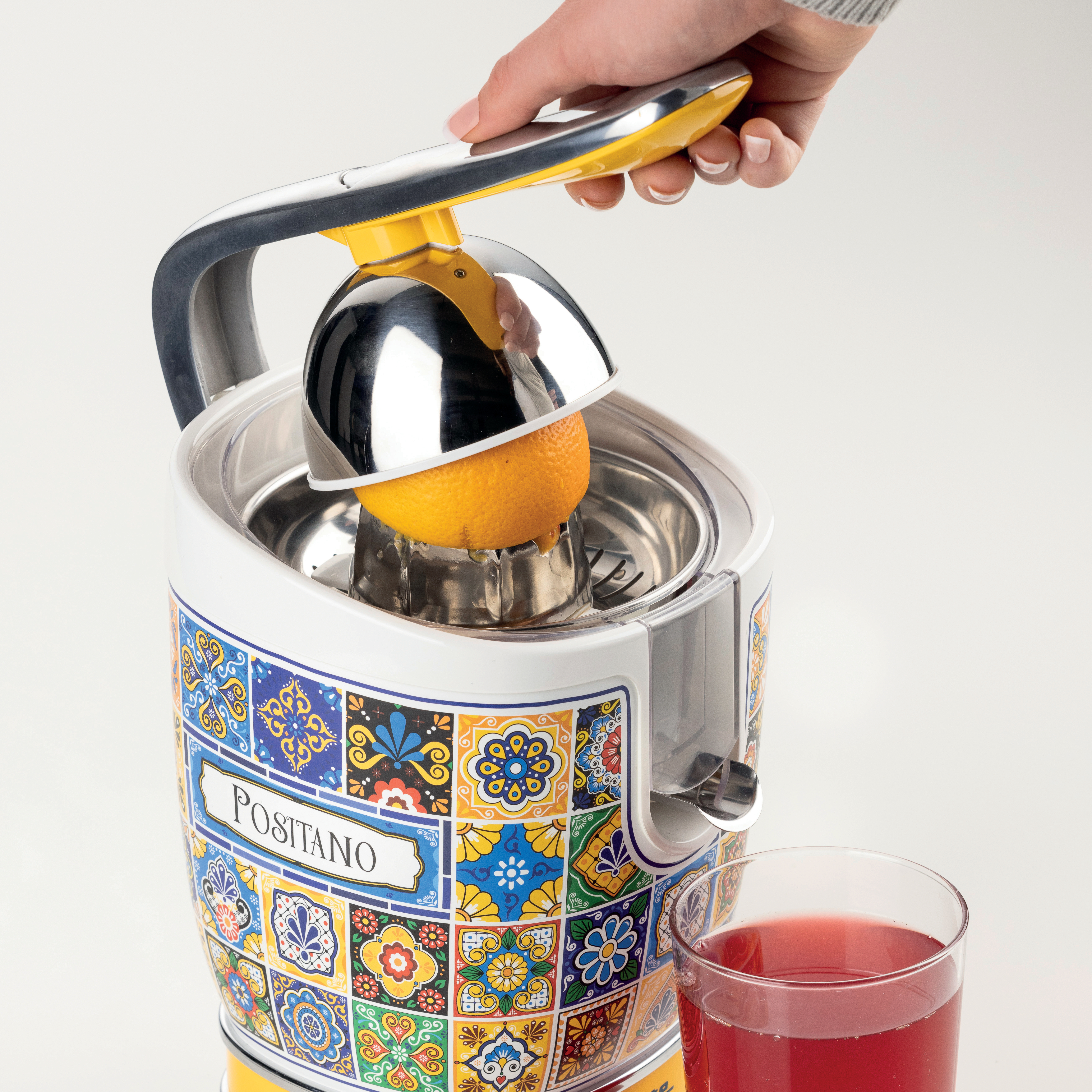 Eine Hand presst eine Orange mit einer Saftpresse aus. Die Saftpresse ist weiß und verziert, und rechts steht ein Glas Saft.